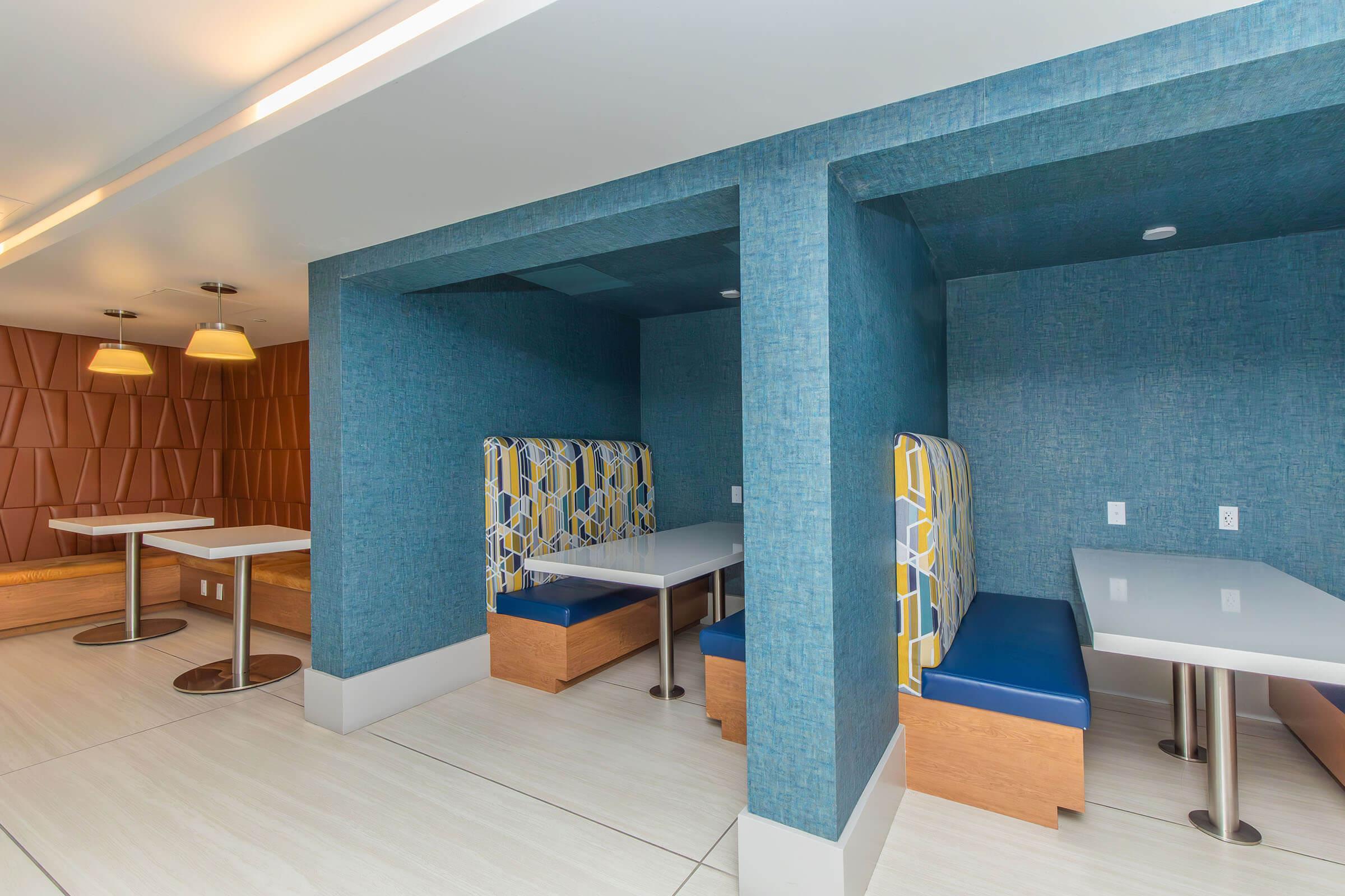 Modern interior featuring two cozy seating booths with blue and patterned upholstery, small white tables, and a textured blue wall. The space is well-lit, with soft lighting fixtures above and a contrasting brown panel wall in the background.