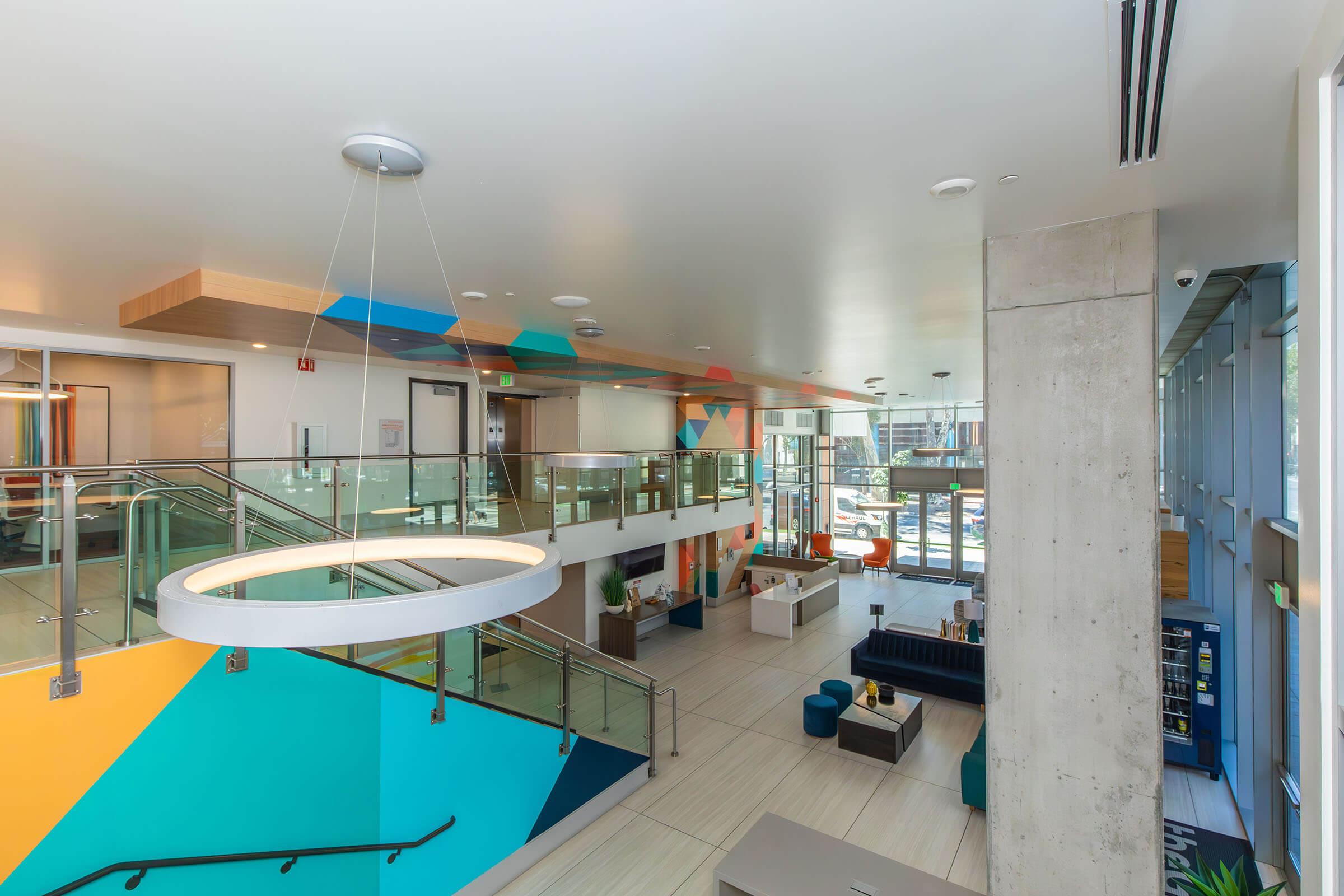 A modern, spacious lobby area featuring a multi-level design. Brightly colored geometric patterns adorn the walls, with stylish furniture such as couches and a reception area. Large windows allow natural light to fill the space, creating an inviting atmosphere. A circular light fixture hangs from the ceiling.