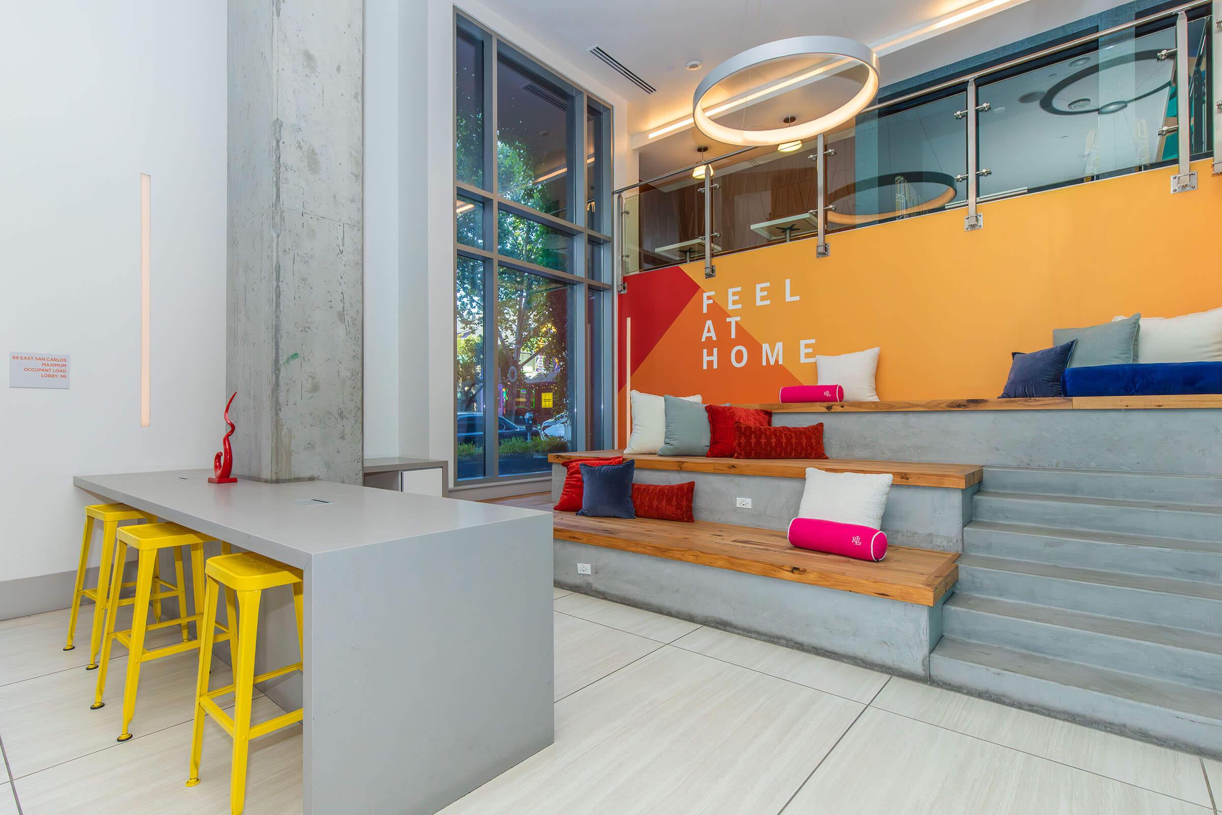 A modern indoor space featuring a colorful wall with the text "FEEL AT HOME." There are steps leading up to a cozy seating area with various cushions in red, blue, and pink. A minimalist gray countertop with yellow stools is located in the foreground, and large windows provide natural light.