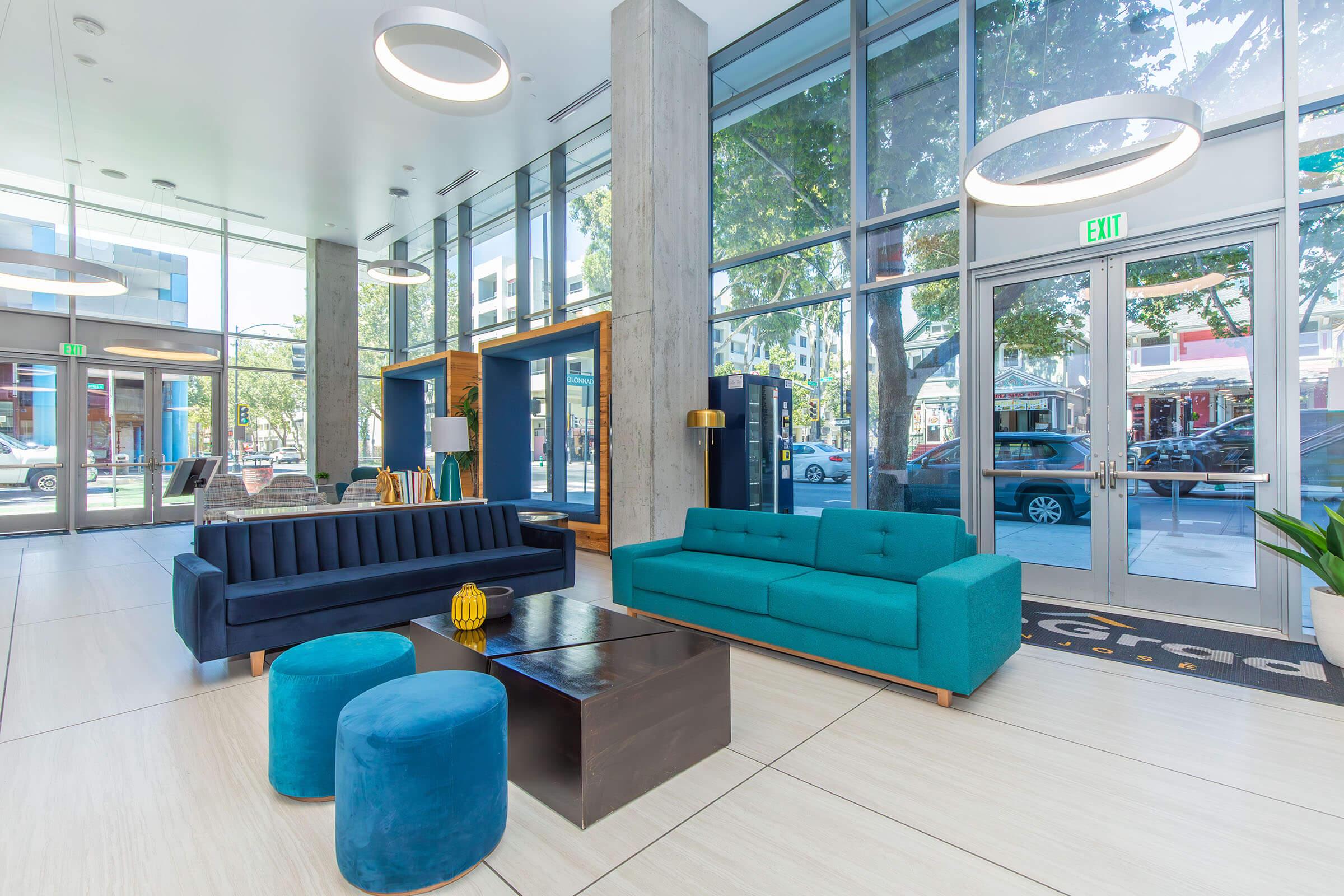 Modern lobby interior featuring large windows, a blue sofa, a teal couch, and a dark coffee table. There are round stools in front of the coffee table, and stylish light fixtures above. The space is bright with natural light and has a contemporary design.
