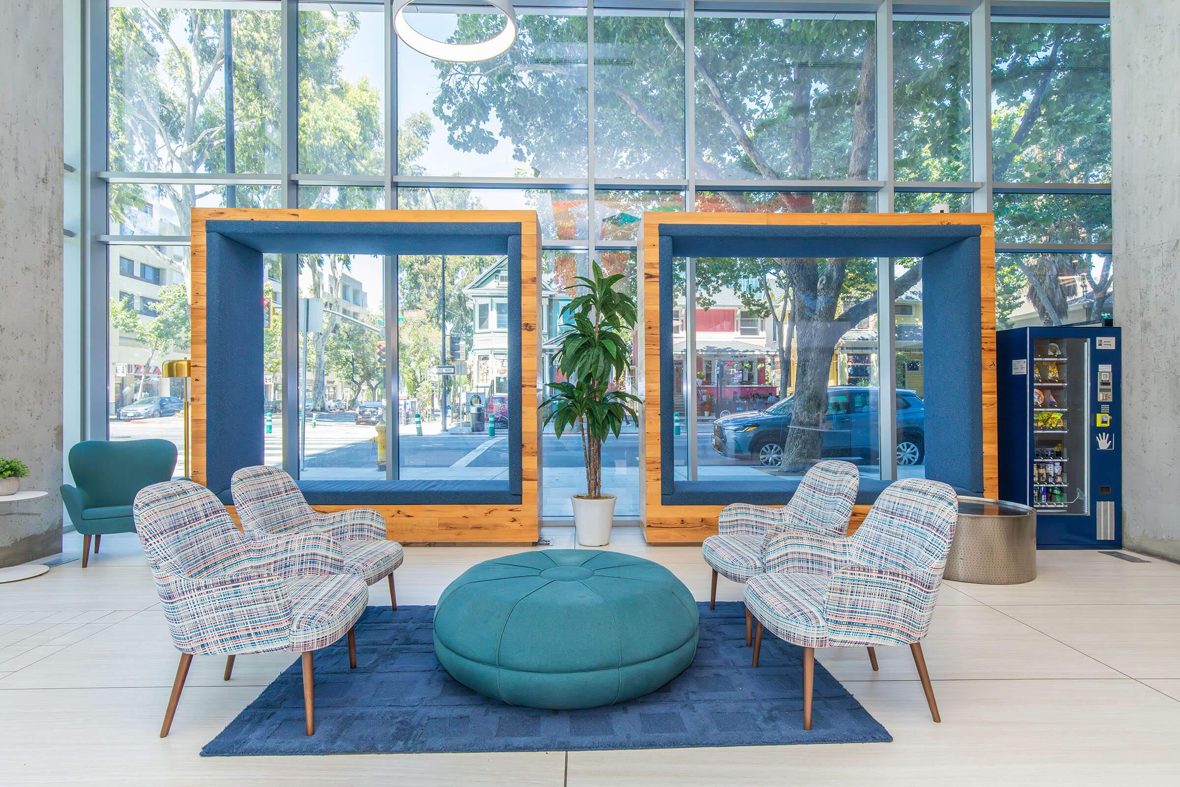 Modern lobby space featuring two large wooden frames, potted plant, and four patterned armchairs surrounding a circular teal ottoman. Floor-to-ceiling windows provide natural light and a view of the street outside. A vending machine is visible in the background.
