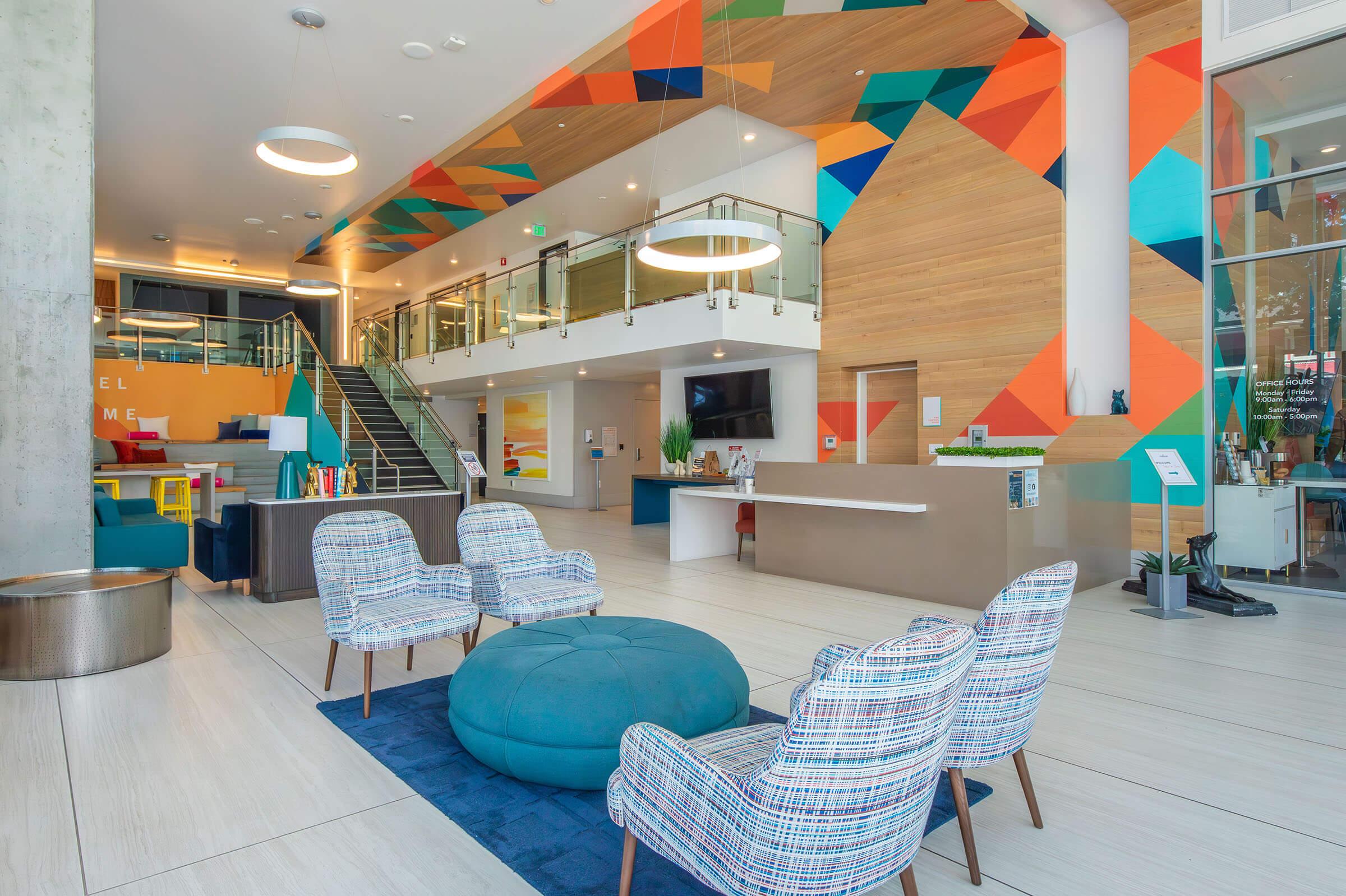 Modern hotel lobby featuring stylish seating with patterned chairs and a large circular ottoman. Brightly colored geometric wall art complements the wooden accents. A reception desk is visible, alongside stairs leading to an upper level, creating a welcoming and contemporary atmosphere.