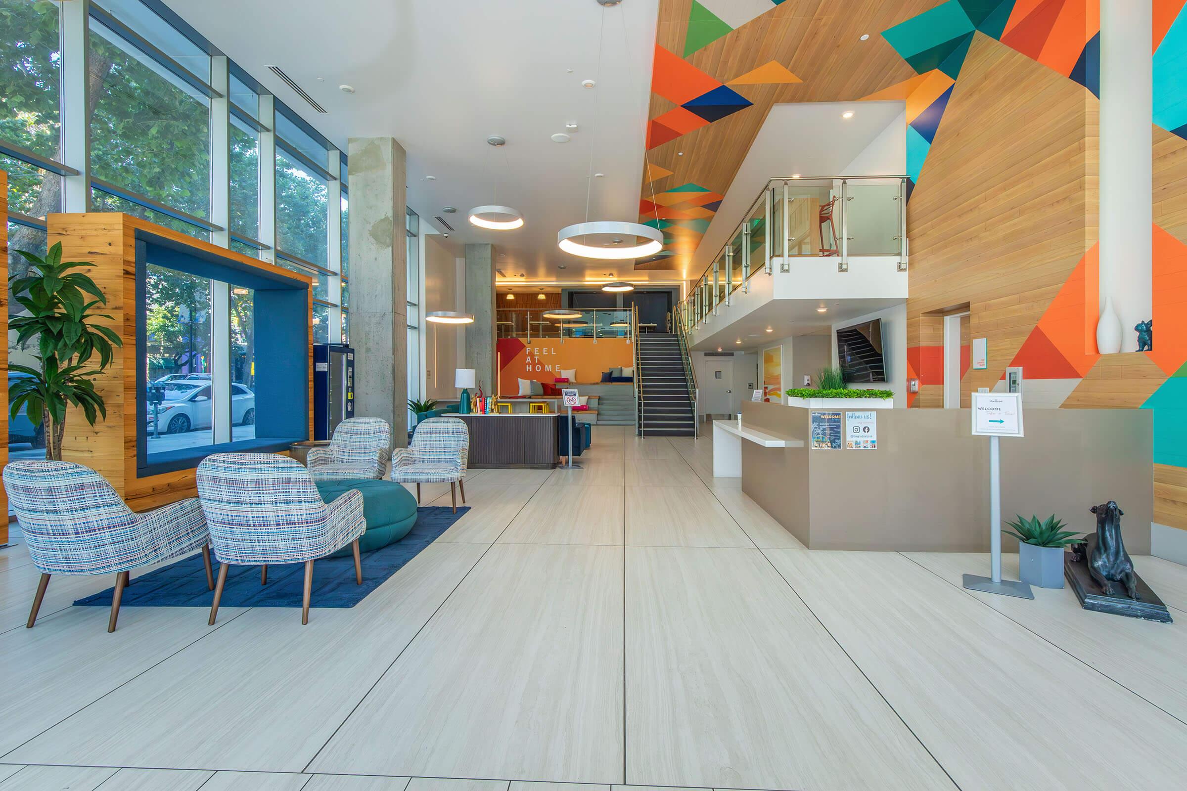 A modern lobby with a spacious design featuring colorful geometric wall art, wooden accents, and comfortable seating areas. There are two patterned chairs and a small green ottoman on the floor. A reception desk is visible, along with a staircase leading to an upper level. Large windows let in natural light.