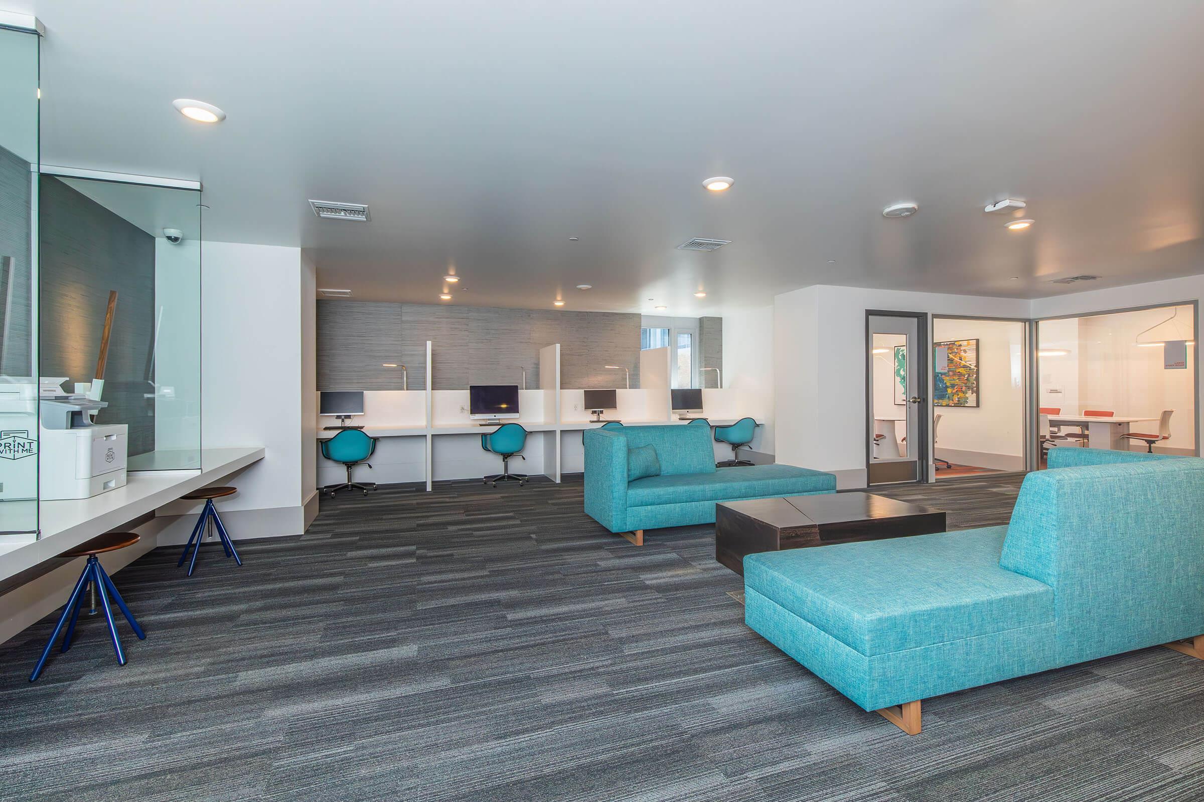 A modern office space featuring stylish blue seating, a coffee table, and workstations with computers. The floor has gray carpeting, and there are glass partitions separating areas. The walls are light-colored, creating a bright and inviting atmosphere.
