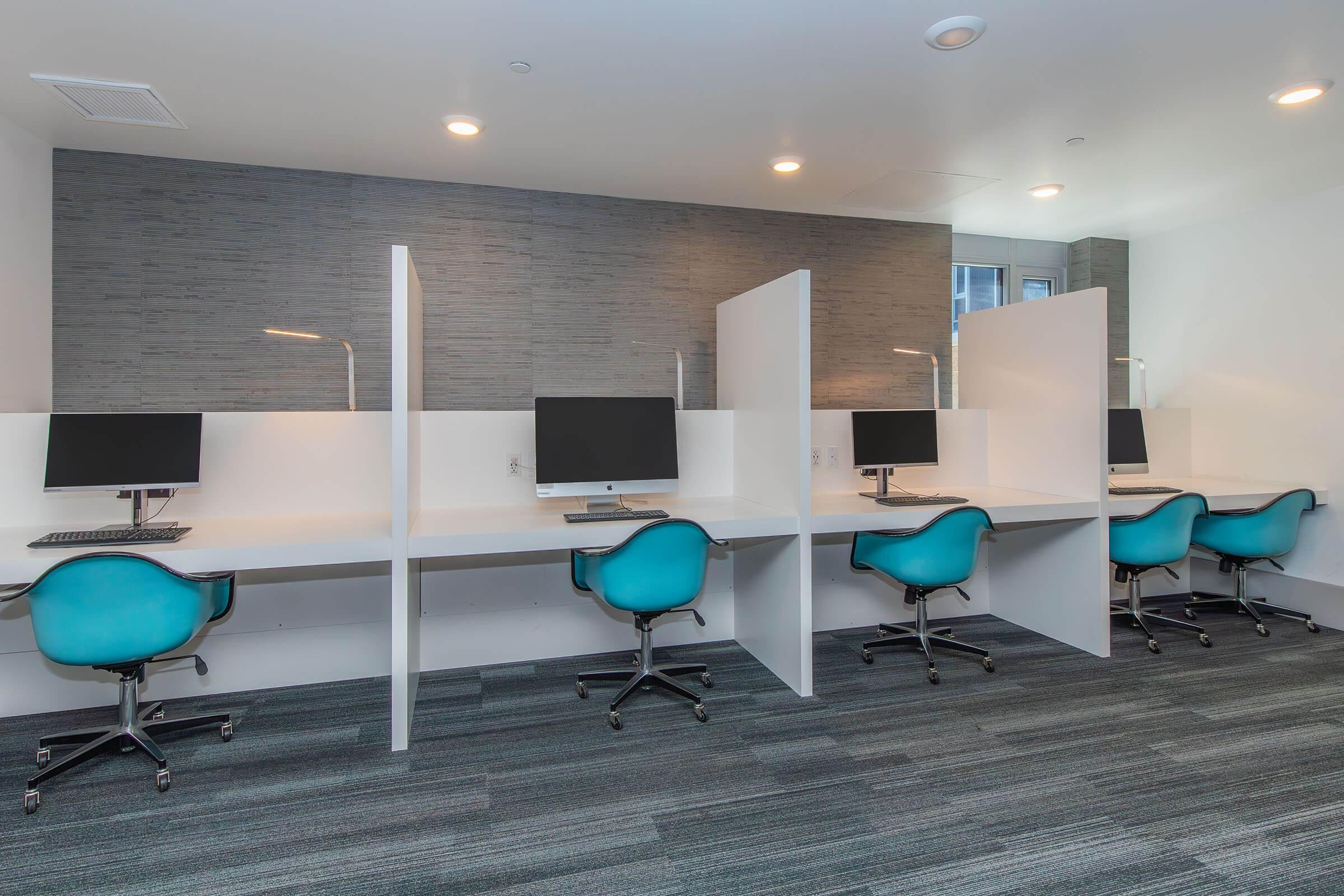 A modern office space featuring four workstations. Each workstation has a sleek white desk and a computer monitor. Teal chairs are positioned in front of the desks, and there are dividers for privacy. The walls are a soft gray, and the space has bright, overhead lighting.