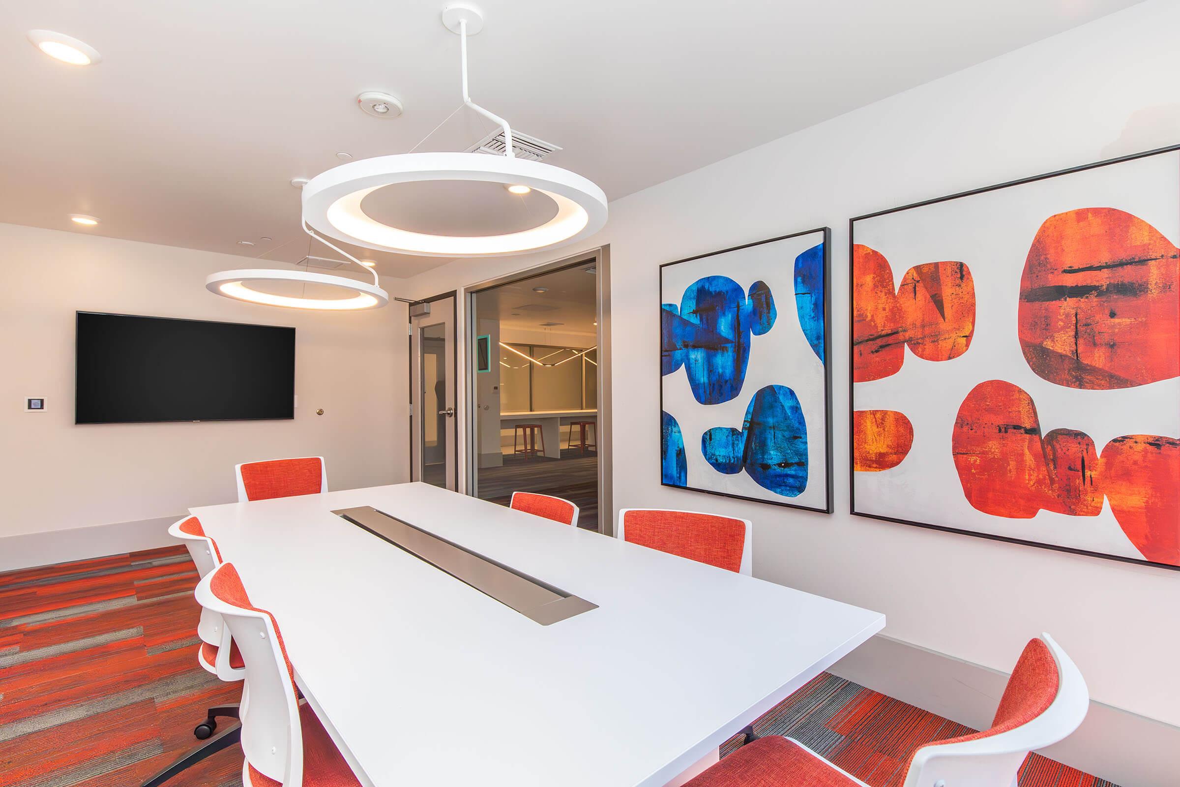 A modern conference room featuring a large white table surrounded by orange-accented chairs. Two abstract art pieces in vibrant red, blue, and orange hues hang on the walls. A flat-screen TV is mounted on one wall, and glass doors lead to another area, with a stylish carpet underfoot.