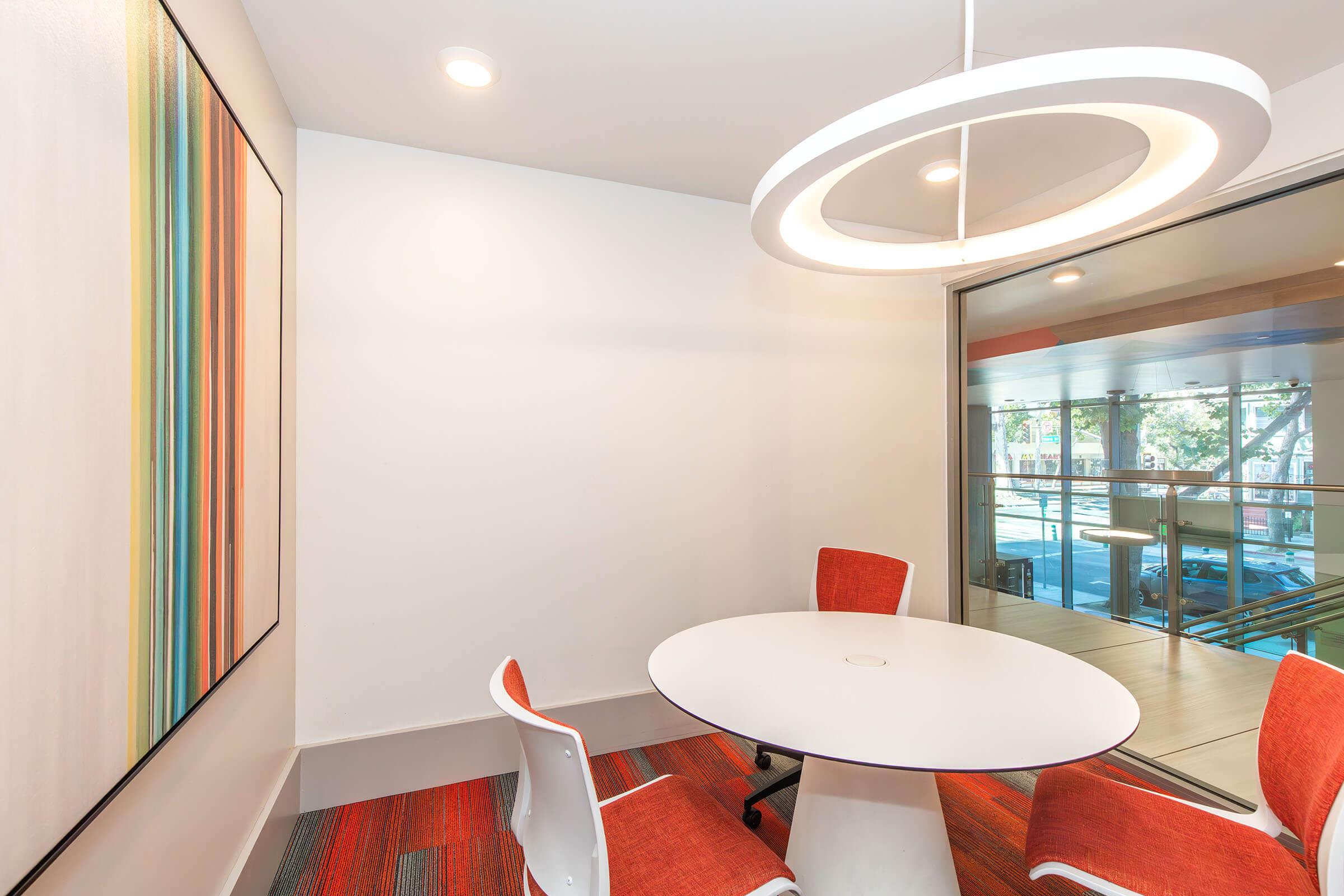 A modern meeting room featuring a round white table surrounded by four orange chairs. One wall showcases a colorful abstract artwork with stripes, while large windows allow natural light to enter the space, creating an open and inviting atmosphere.