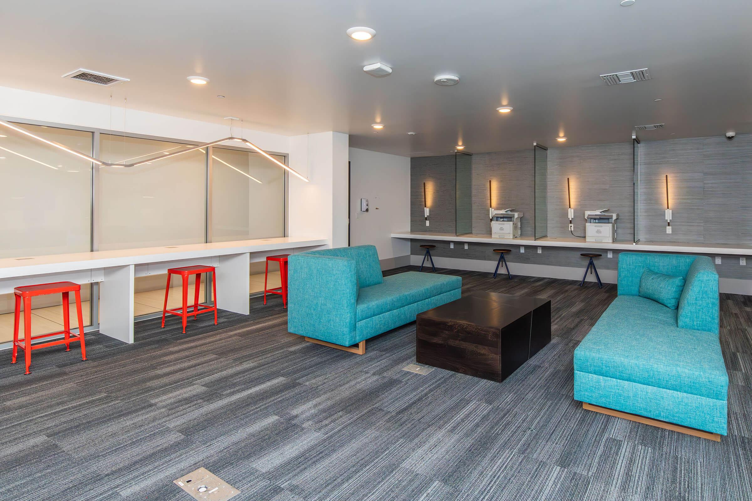 Modern lounge area featuring two teal sofas, a dark coffee table, and bright red stools. There is a long shared workspace desk against the wall with two printers visible in the background. The floor is covered with gray carpeting, and the space has ambient lighting, creating a contemporary and inviting atmosphere.