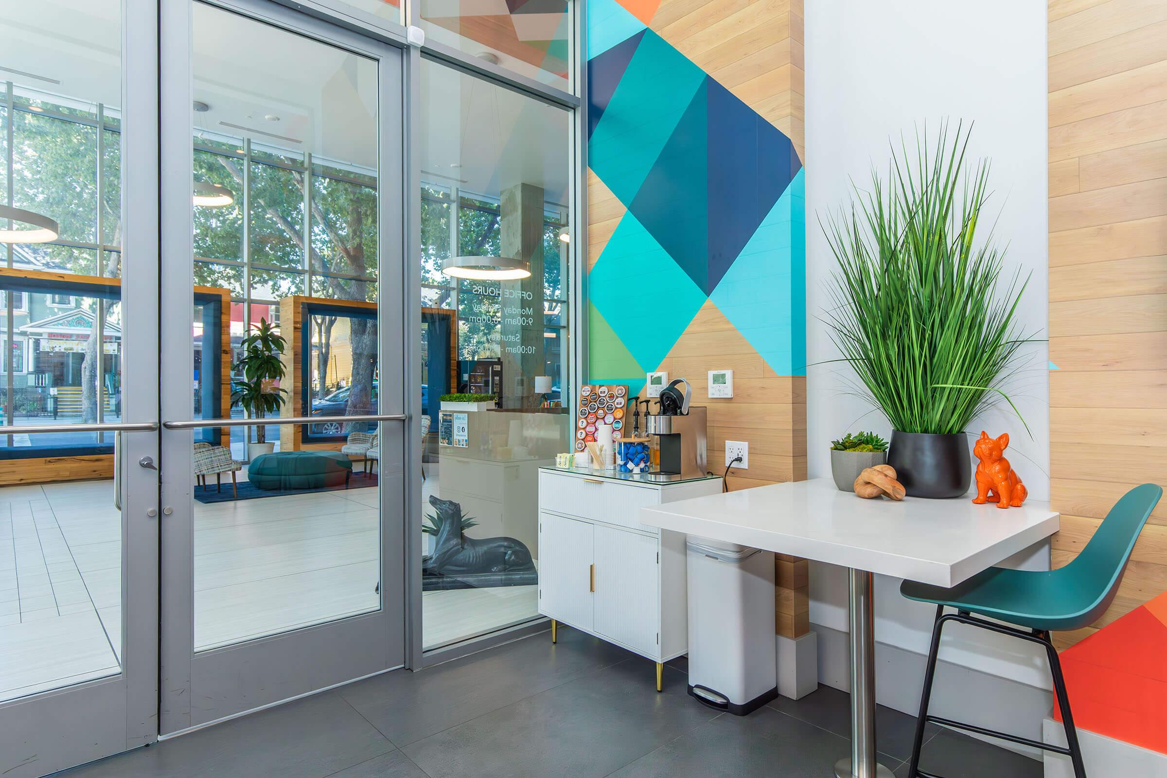 A modern entryway featuring glass doors, a vibrant geometric wall design, and a small white table with a coffee maker and decorative items. There is a green plant and an orange decorative fox on the table, accompanied by a green chair. The area is well-lit with natural light and has a contemporary feel.