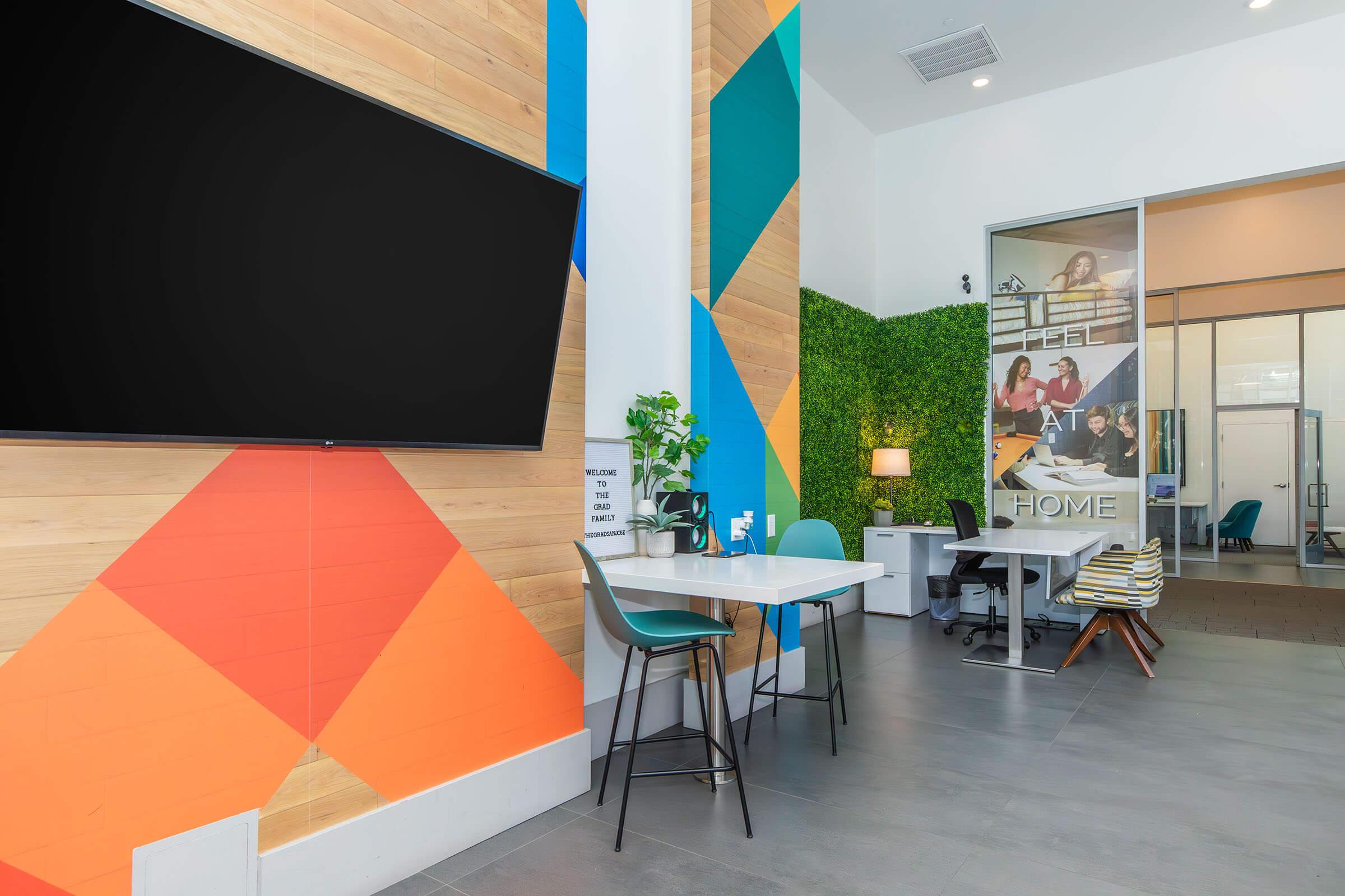 Modern office space featuring colorful geometric wall designs, a large TV screen, a table with chairs, and a green plant. The room includes workspaces with a desk and computer, alongside a decorative sign promoting a welcoming atmosphere. Natural light enhances the vibrant interior.