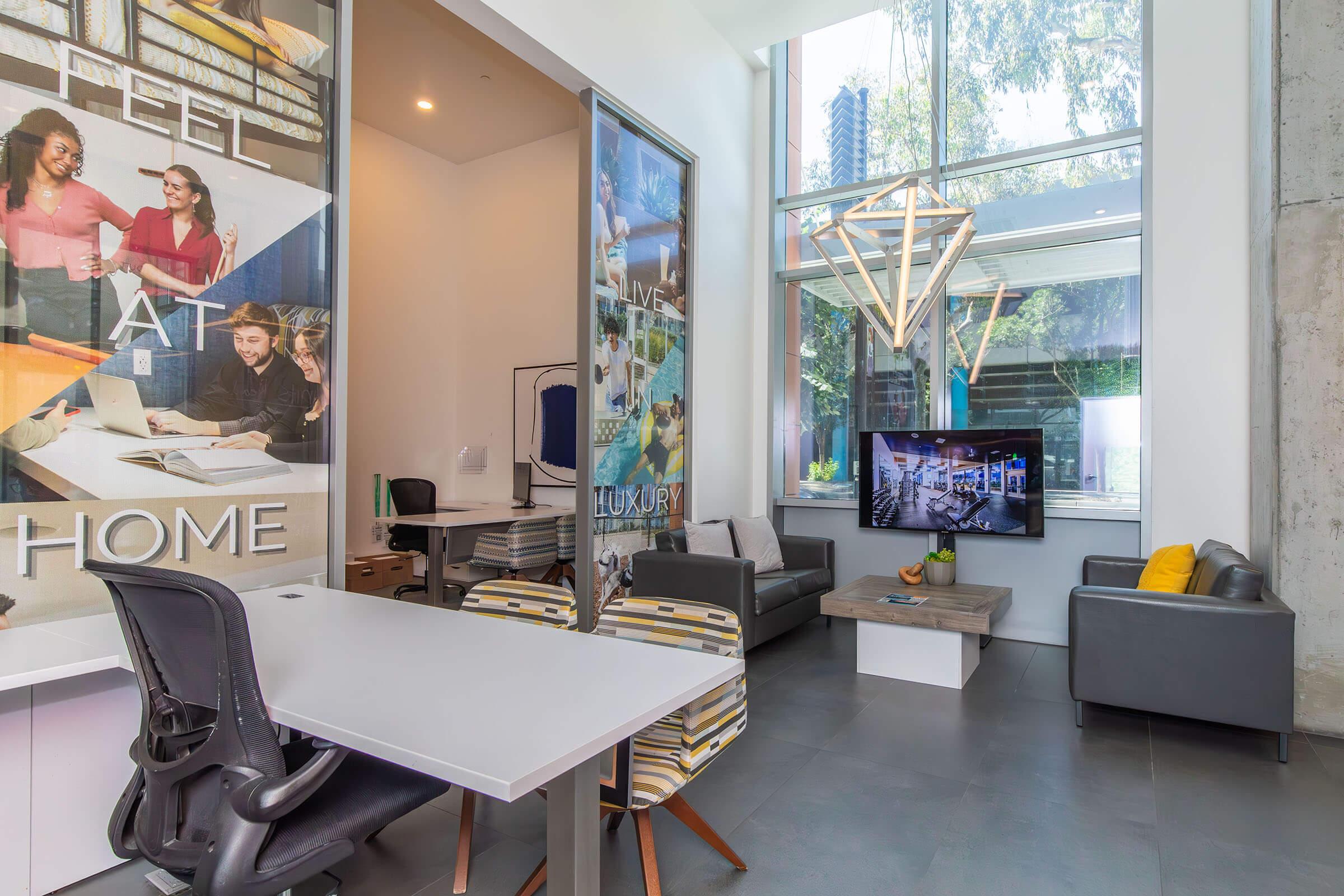 A modern interior space featuring a cozy seating area with a sofa and coffee table, a flat-screen TV on the wall, and large windows allowing natural light. There are stylish design elements, including a geometric light fixture, and a desk with a chair. Decor includes motivational phrases and vibrant artwork.