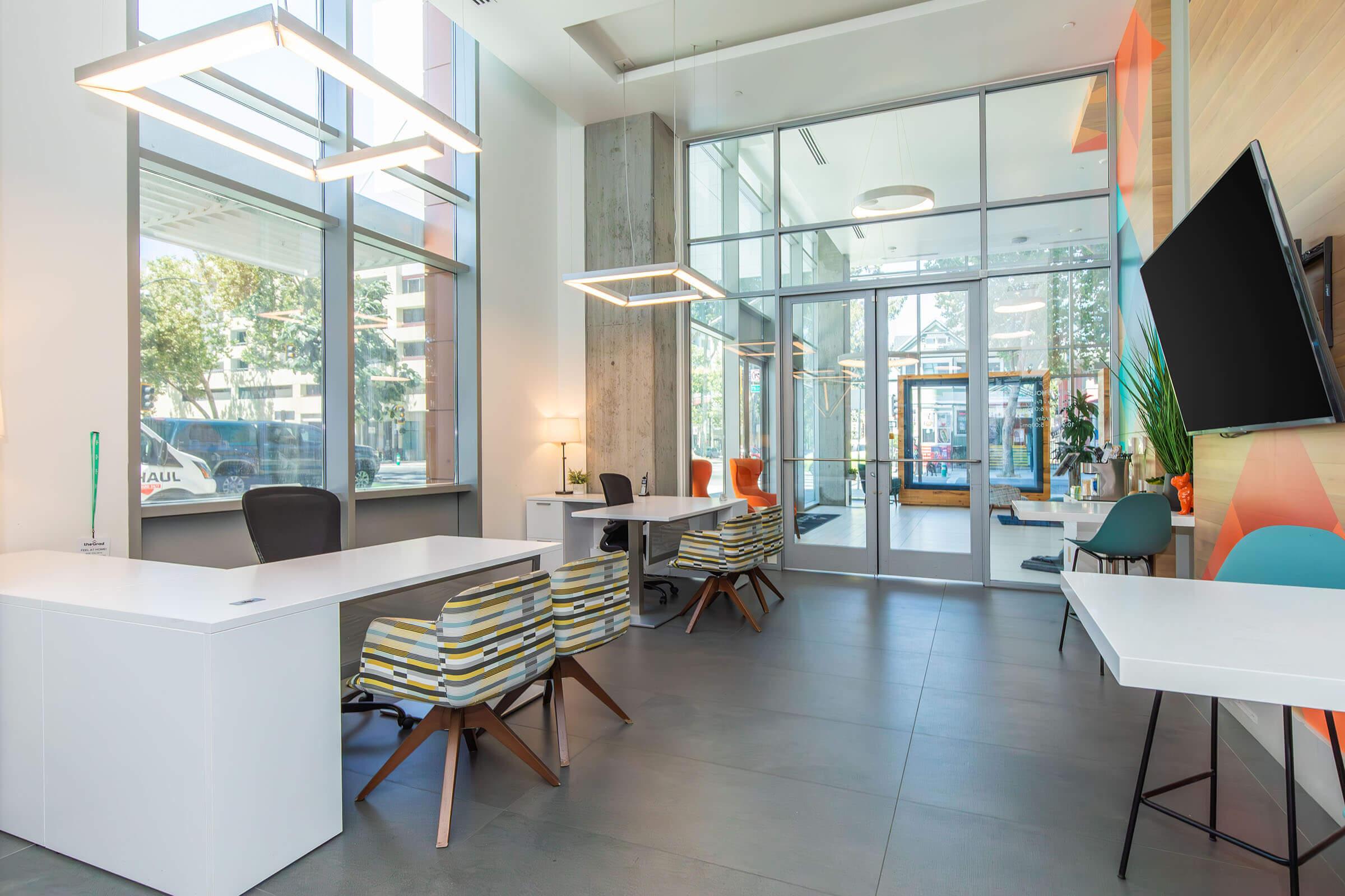 A modern office space featuring large windows, contemporary furniture, and a bright atmosphere. Desks are arranged with stylish chairs in various colors. The entrance shows outdoor greenery, and a flat-screen TV is mounted on the wall, creating a welcoming and functional work environment.