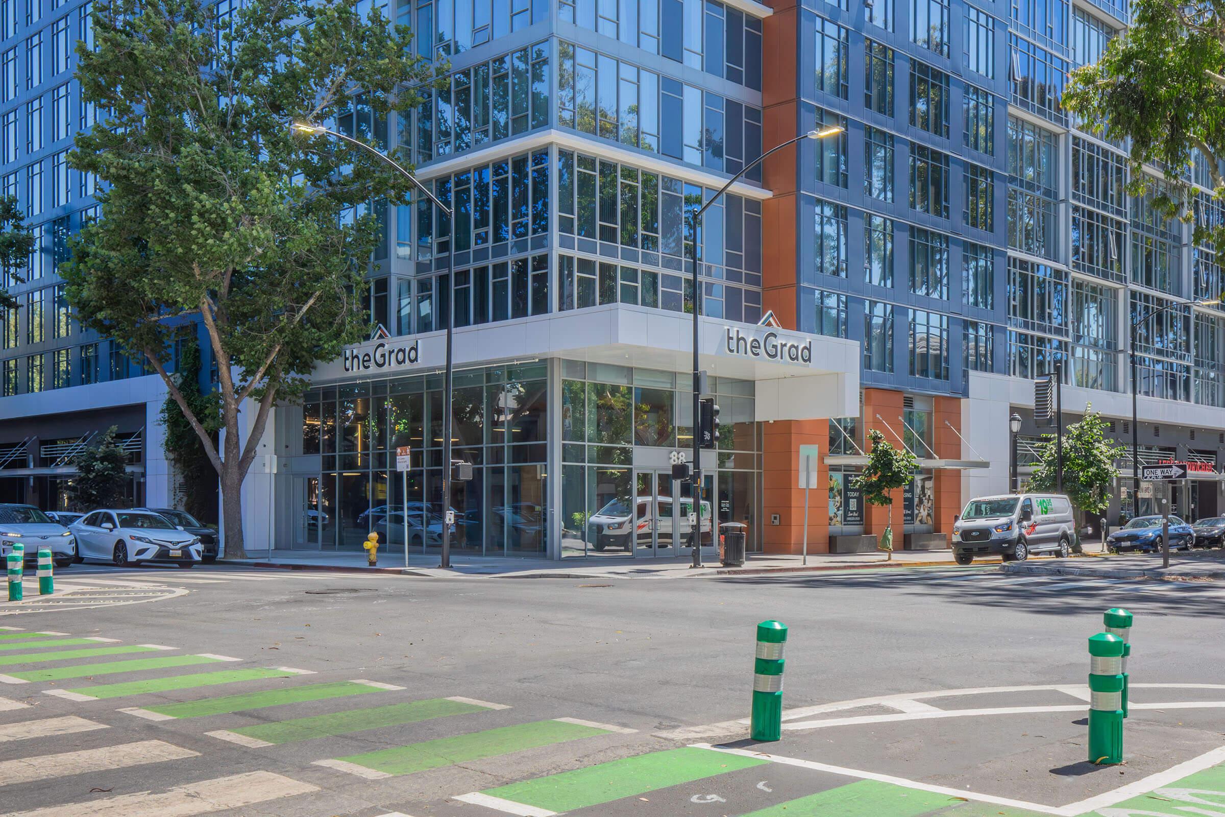 A modern building featuring large glass windows and a storefront labeled "theGrad." The structure is at a street corner with green bicycle lanes and green-and-white striped traffic posts. Surrounding trees provide shade on a sunny day, with vehicles parked nearby.