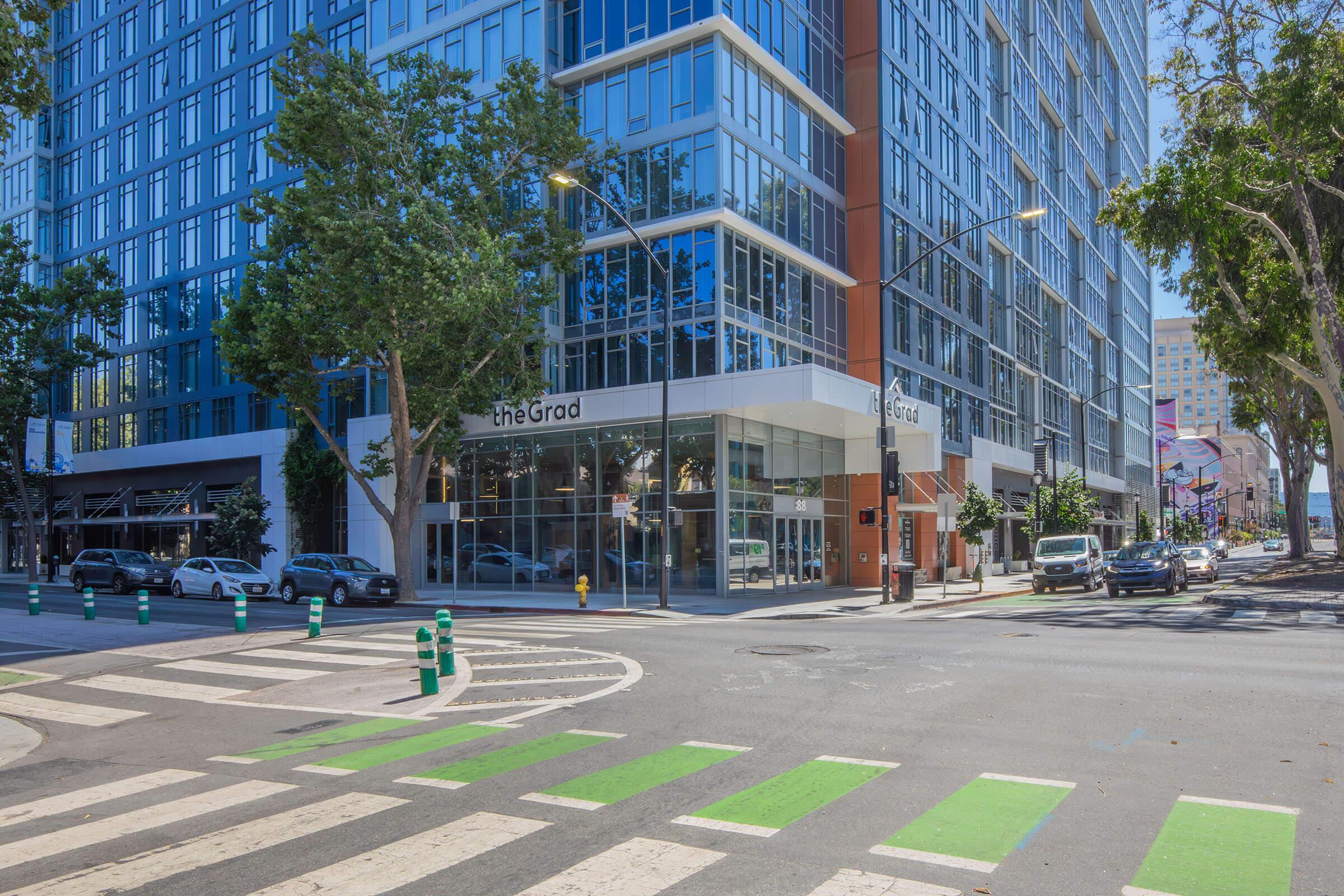 A modern building with large glass windows, featuring the sign "theGrad" at the entrance. The street in front has a crosswalk with green stripes, surrounded by trees and parked cars. The scene is bright and sunny, highlighting an urban environment.