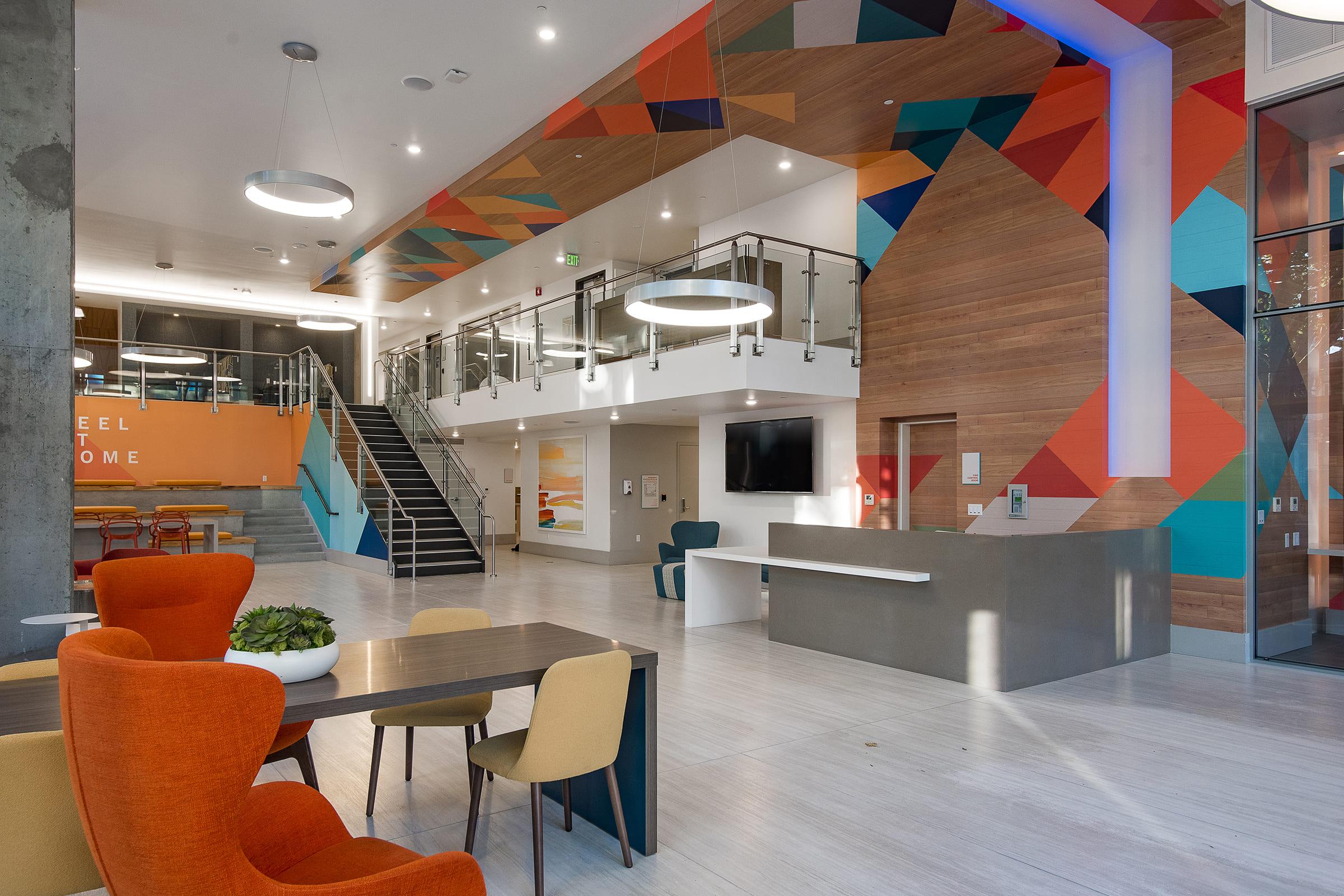 A modern lobby interior featuring a spacious layout with colorful geometric wall designs. There are comfortable seating areas with orange and yellow chairs, a wooden reception desk, a large flat-screen TV on the wall, and a staircase leading to an upper level. Bright lighting enhances the vibrant atmosphere.