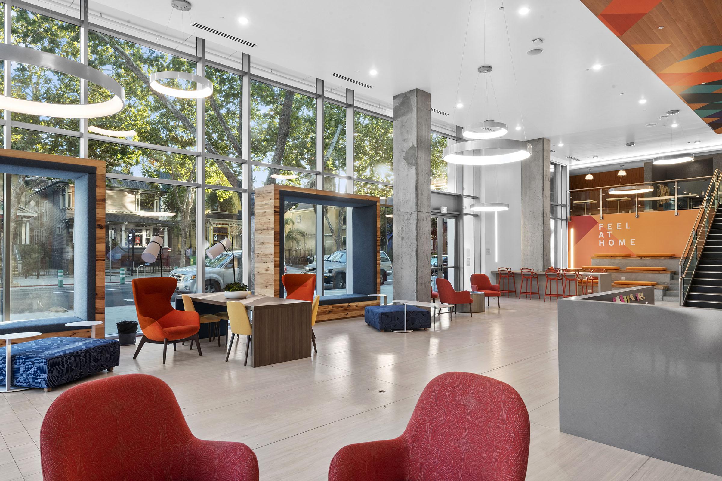 A modern lobby with large windows allowing natural light to flow in. Stylish red and blue chairs are arranged in a welcoming layout. A reception desk is visible, along with a wall featuring the phrase "FEEL AT HOME." The overall atmosphere is bright and inviting, perfect for guests.