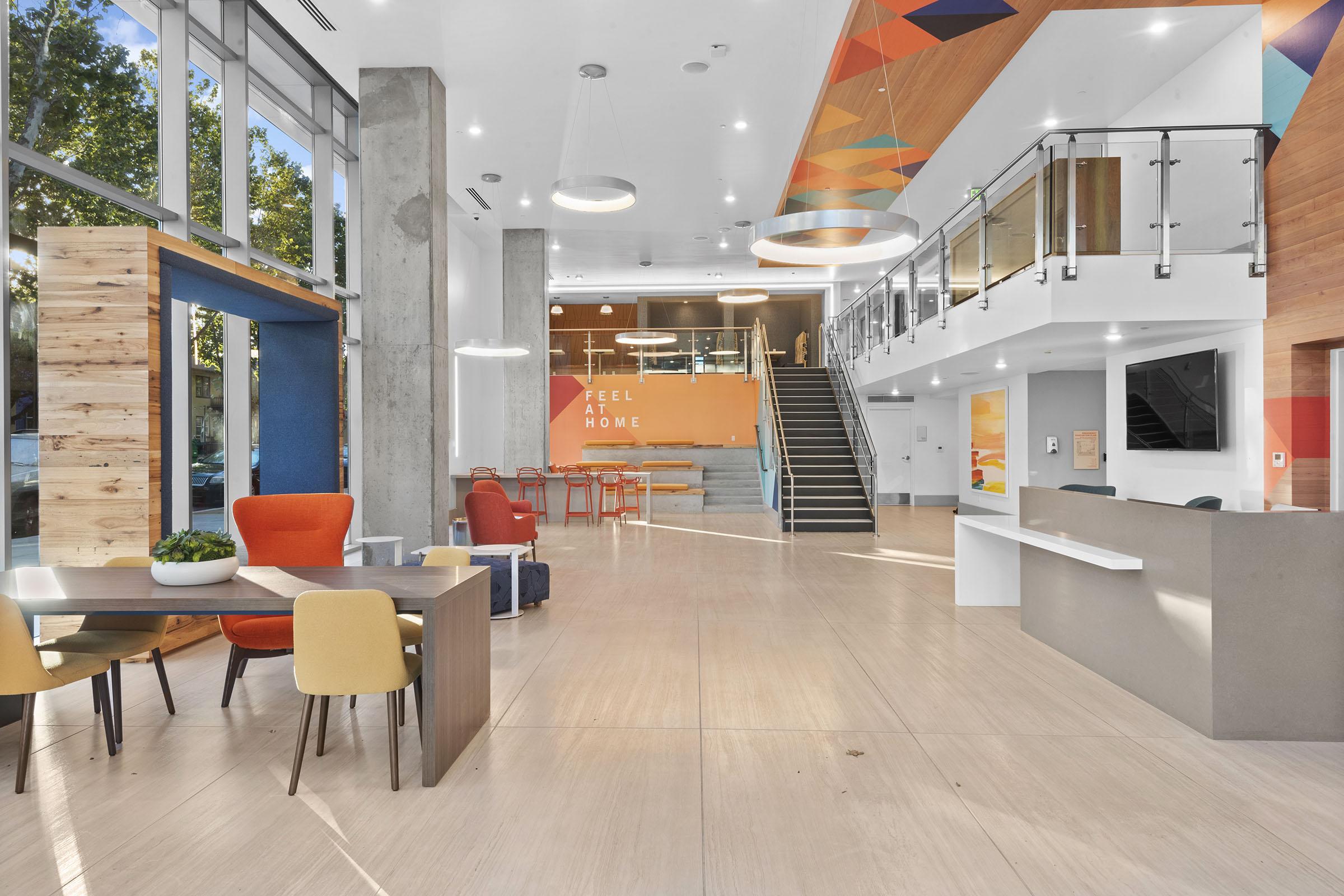 Modern lobby space featuring a reception desk, stylish seating areas with orange and blue chairs, wooden accents, and vibrant wall decorations. Stairs lead to an upper level, and large windows allow natural light to fill the area. The atmosphere is welcoming and contemporary, ideal for visitors.