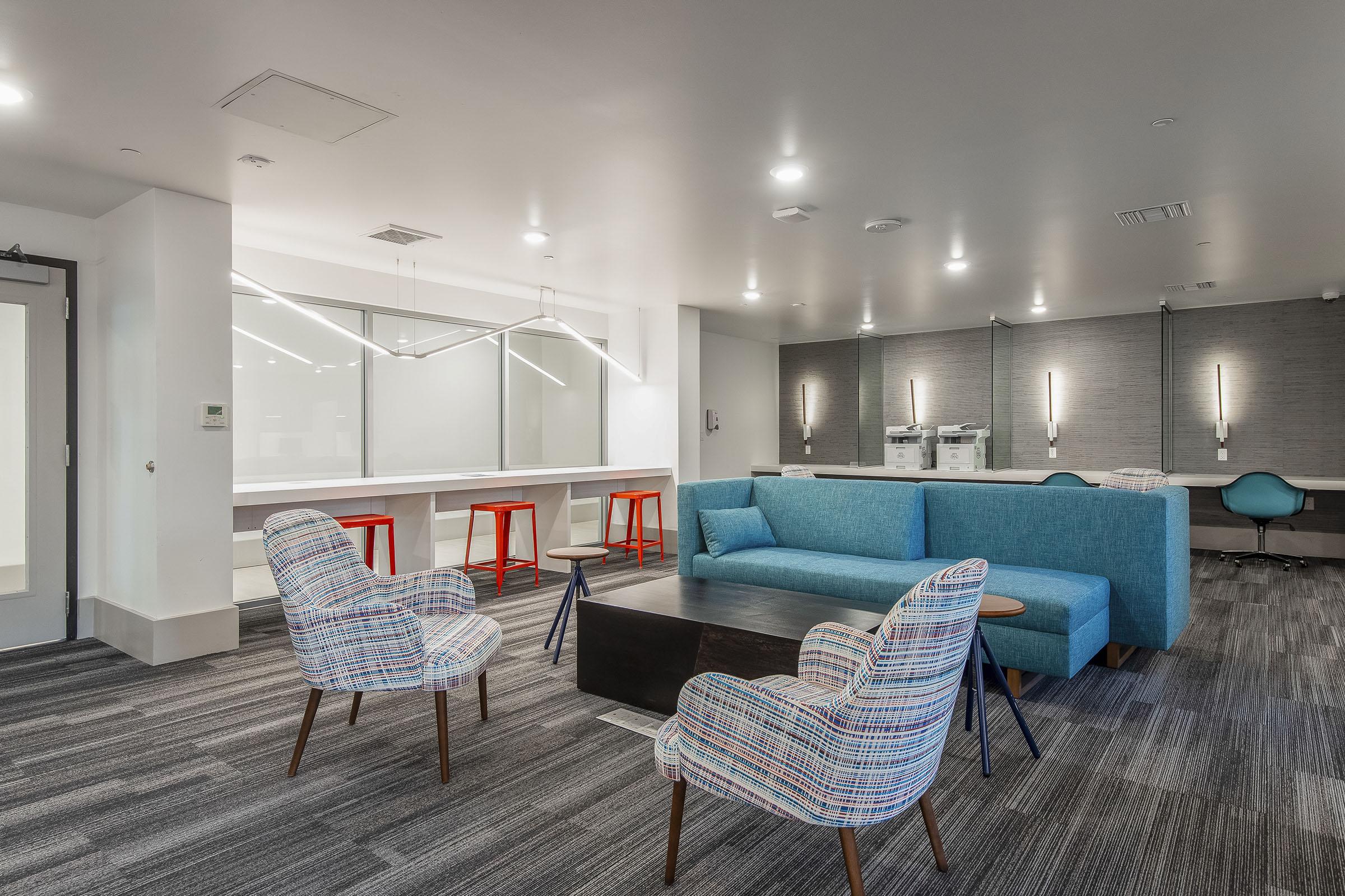 A modern lounge area featuring a blue couch, two patterned armchairs, and a dark coffee table. Red barstools are stationed around a counter with a light gray backdrop. The space is well-lit, showcasing a minimalist design with a blend of soft seating and functional elements.