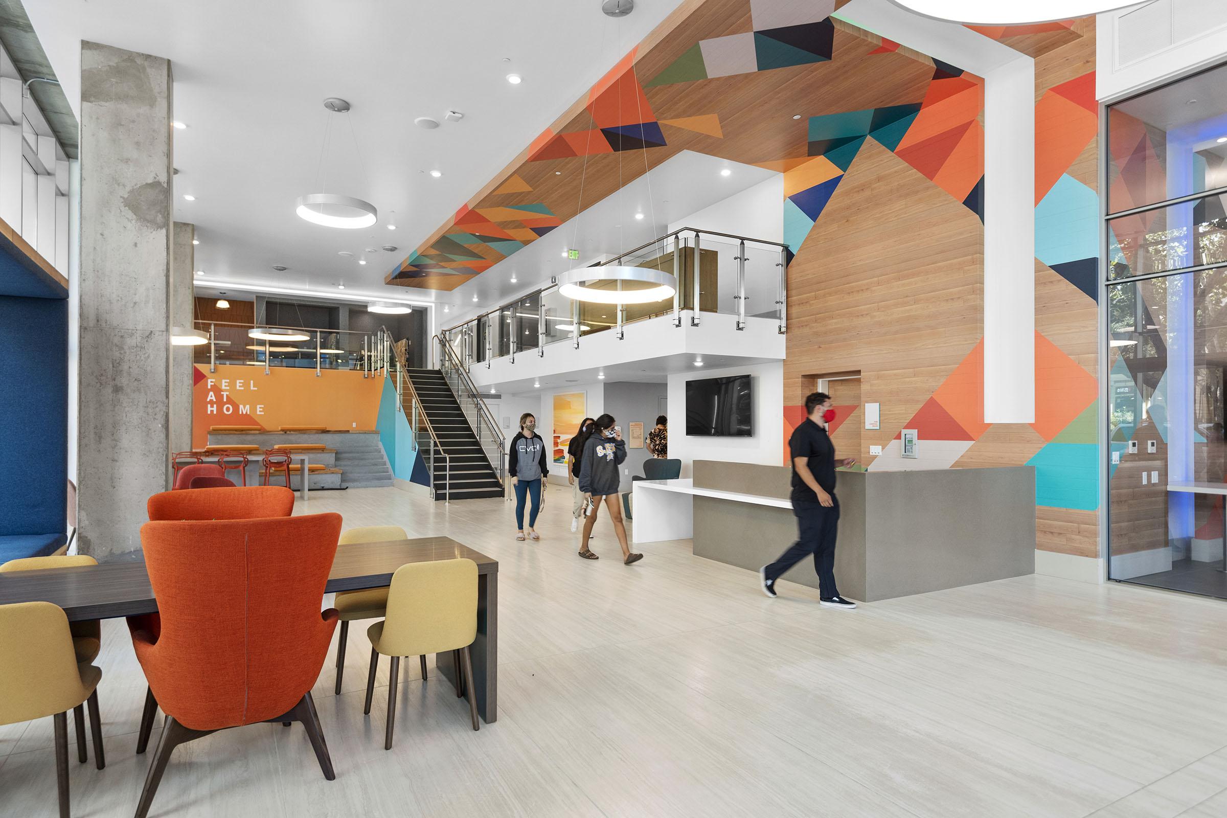 Modern communal space featuring vibrant geometric wall art, a welcoming reception area with a desk, and various seating options. People walk through the area, which includes a staircase leading to an upper level. Large windows provide natural light, creating an inviting atmosphere. 