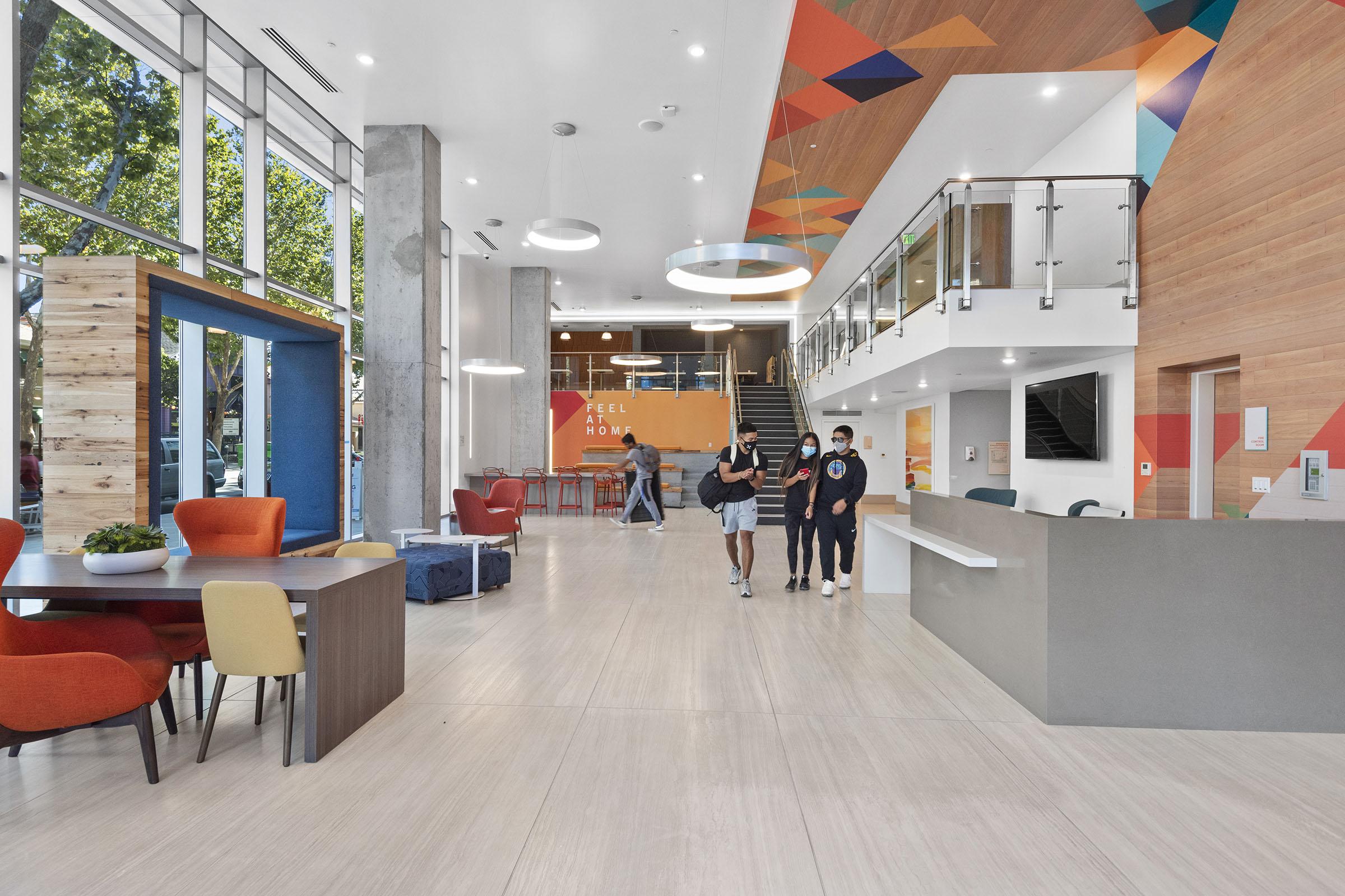 Modern lobby interior featuring a welcoming reception desk, colorful seating areas, and large windows letting in natural light. People walk through the space, which has vibrant wall designs and a contemporary aesthetic. A communal atmosphere is highlighted with an open layout and stylish furniture.