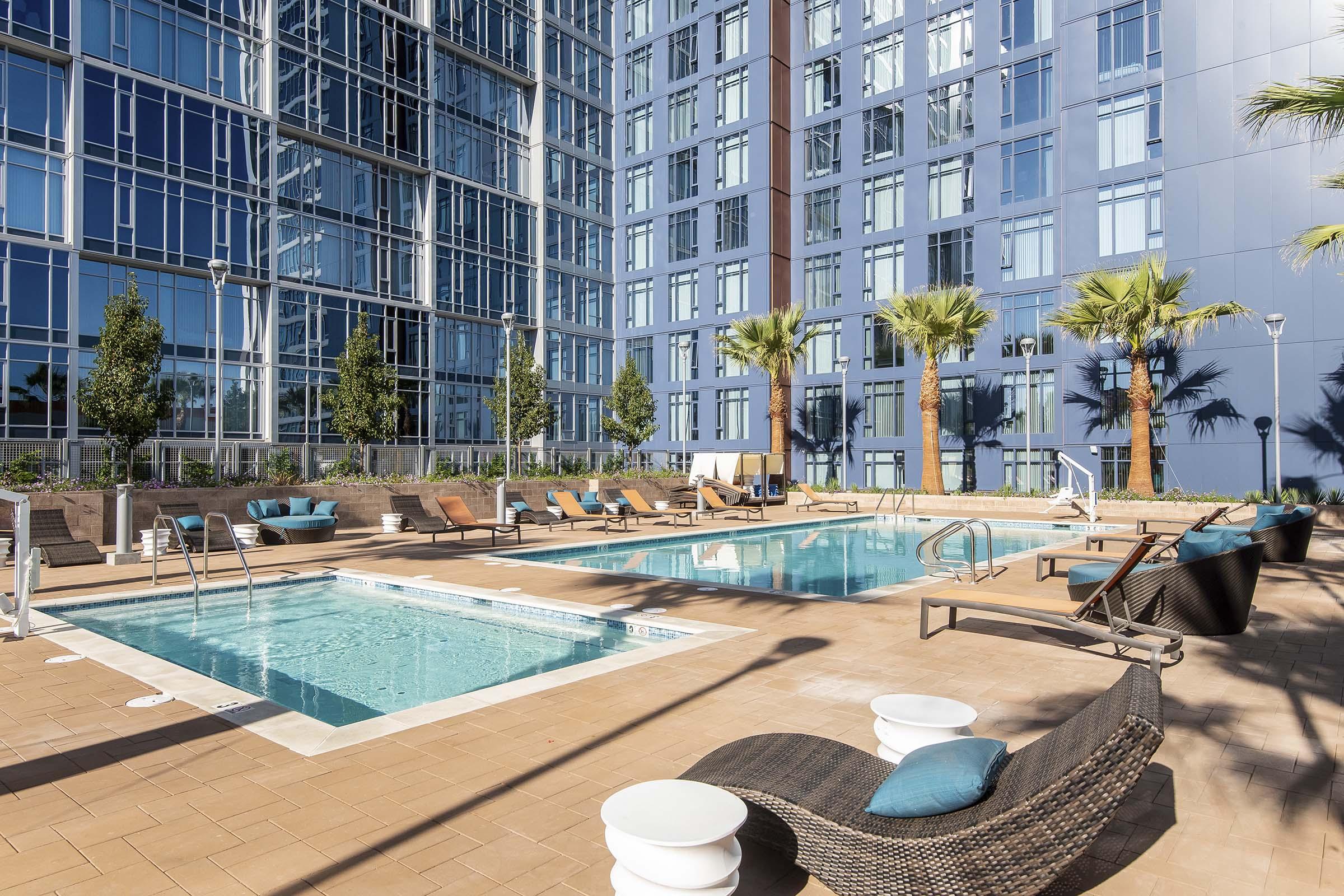 A modern swimming pool area featuring two pools surrounded by palm trees and lounge chairs. The pools are set against a backdrop of a sleek, glass-paneled building, creating a vibrant outdoor space for relaxation and recreation. Sunlight reflects off the water, enhancing the inviting atmosphere.