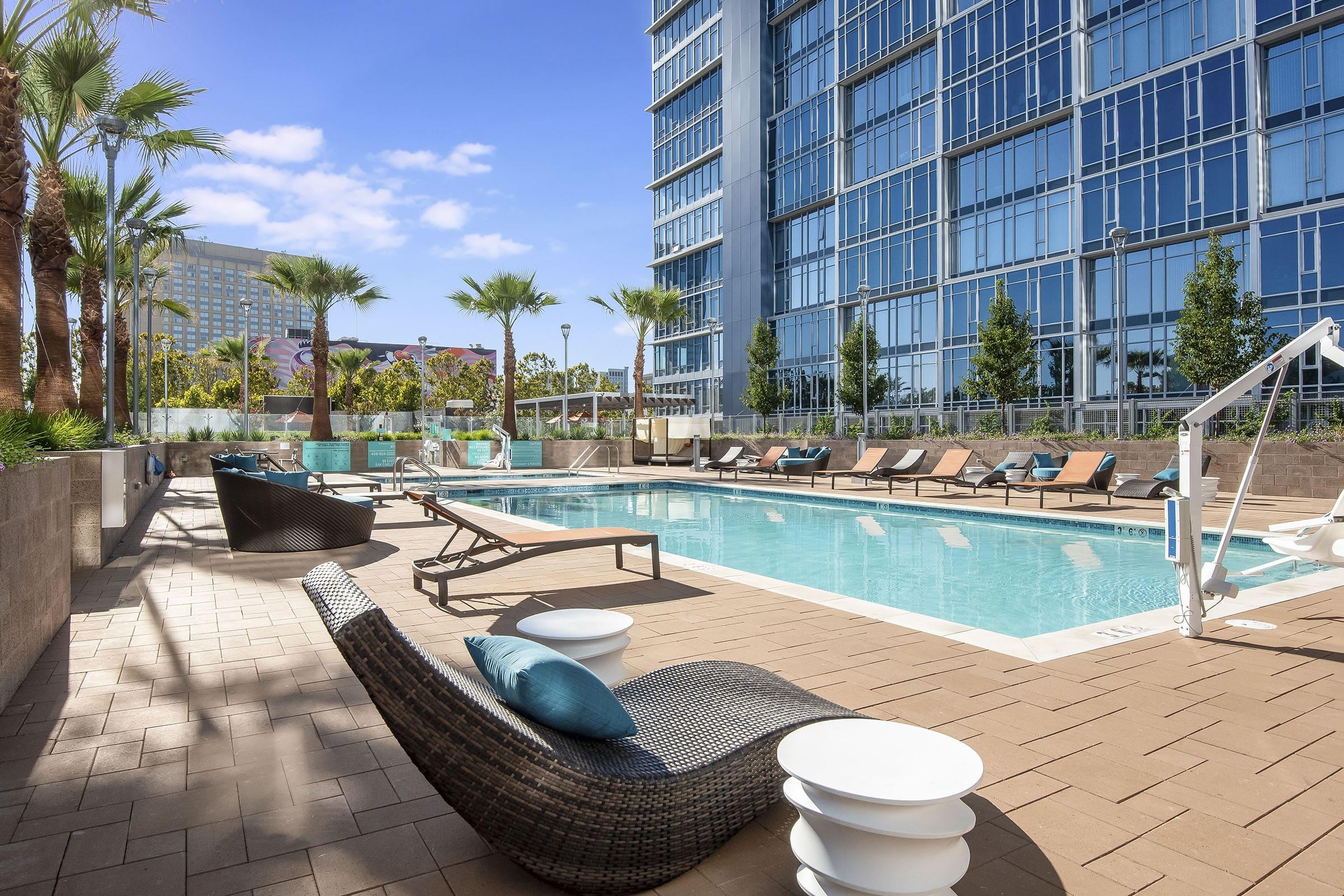 A sunlit outdoor pool area featuring lounge chairs and modern seating. Palm trees line the space, and a high-rise building with glass windows reflects the blue sky in the background. The pool is inviting, surrounded by a stone patio and greenery.