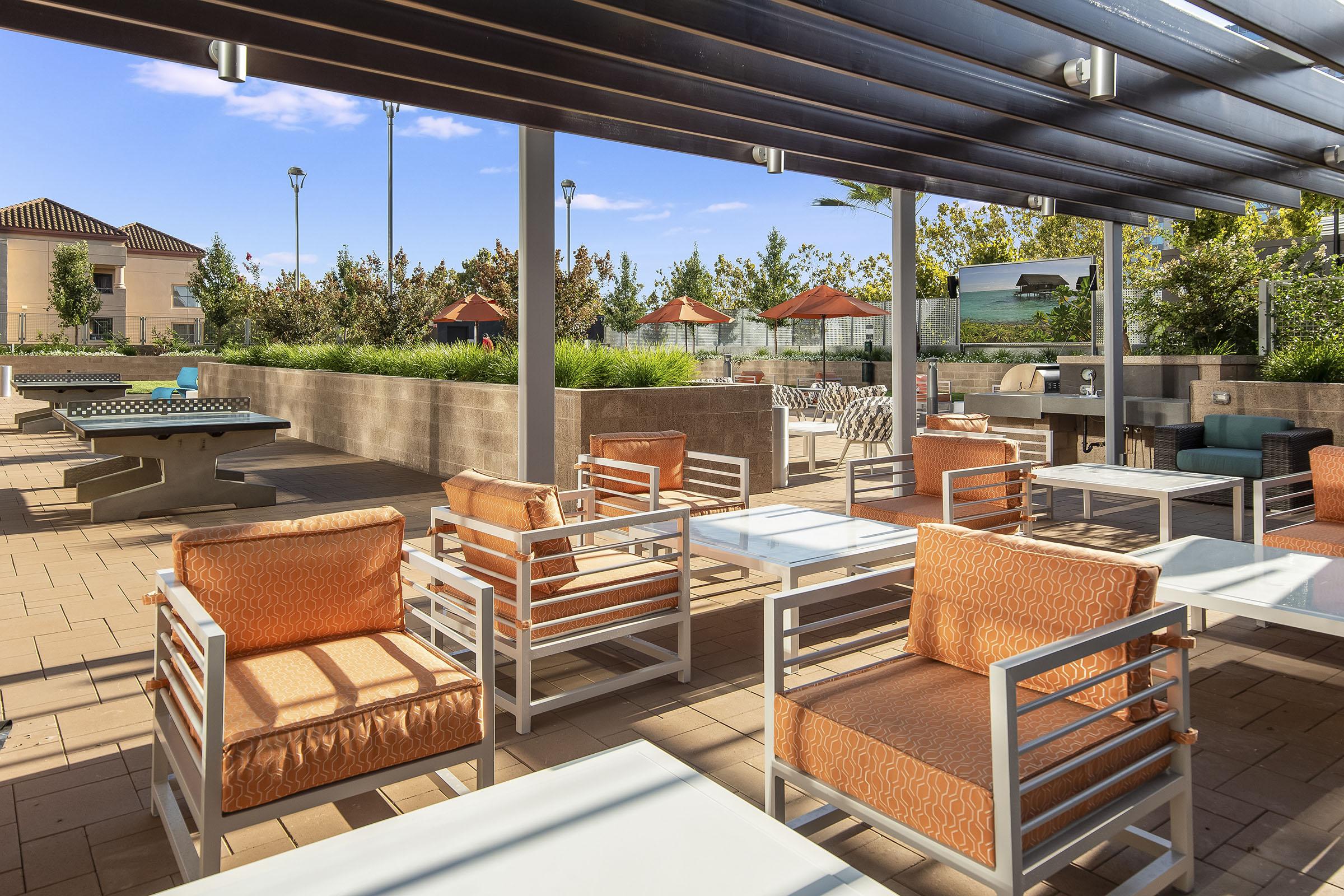 Modern outdoor lounge area featuring comfortable orange upholstered chairs, sleek white tables, and shaded seating under a pergola. Lush greenery and patio umbrellas create a relaxing atmosphere, with additional seating arrangements visible in the background. Sunlight brightens the scene, enhancing the inviting ambiance.