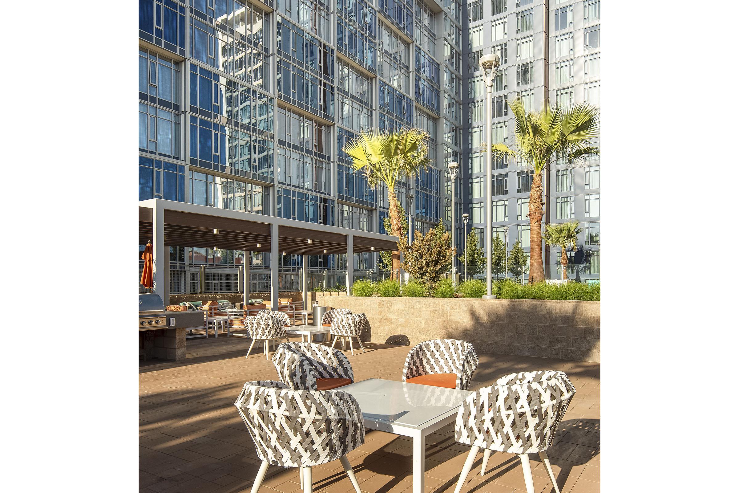 Outdoor seating area with modern furniture, including patterned chairs and white tables, set against a backdrop of a glass-walled building. Lush palm trees and green landscaping enhance the ambiance, creating a stylish and inviting atmosphere.