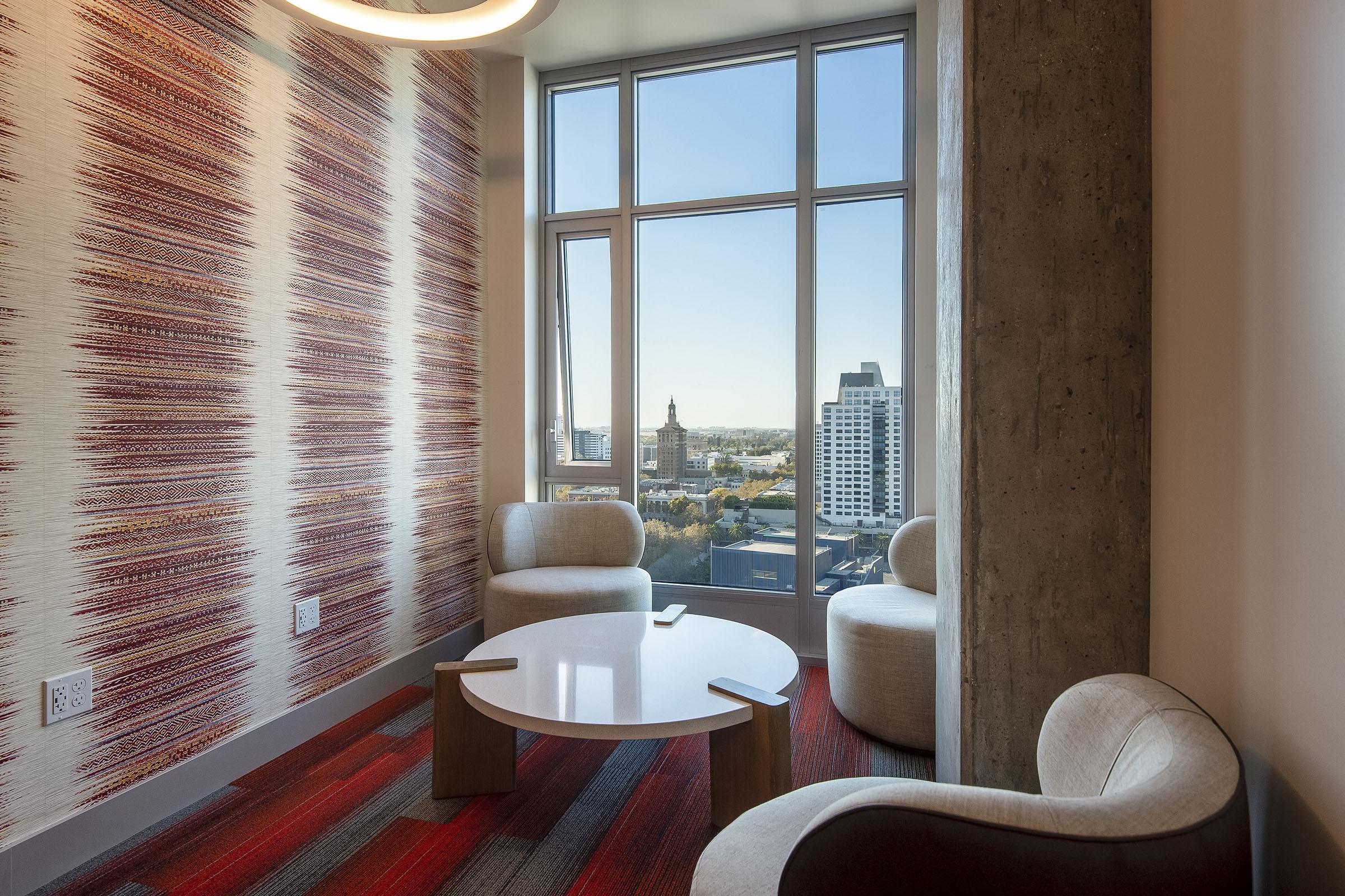 A modern seating area in a well-lit room with large windows showcasing a cityscape. The interior features a striking patterned wall, a round table, and three comfortable chairs, all set on a patterned carpet with shades of red and gray. Natural light illuminates the space, highlighting the contemporary design.