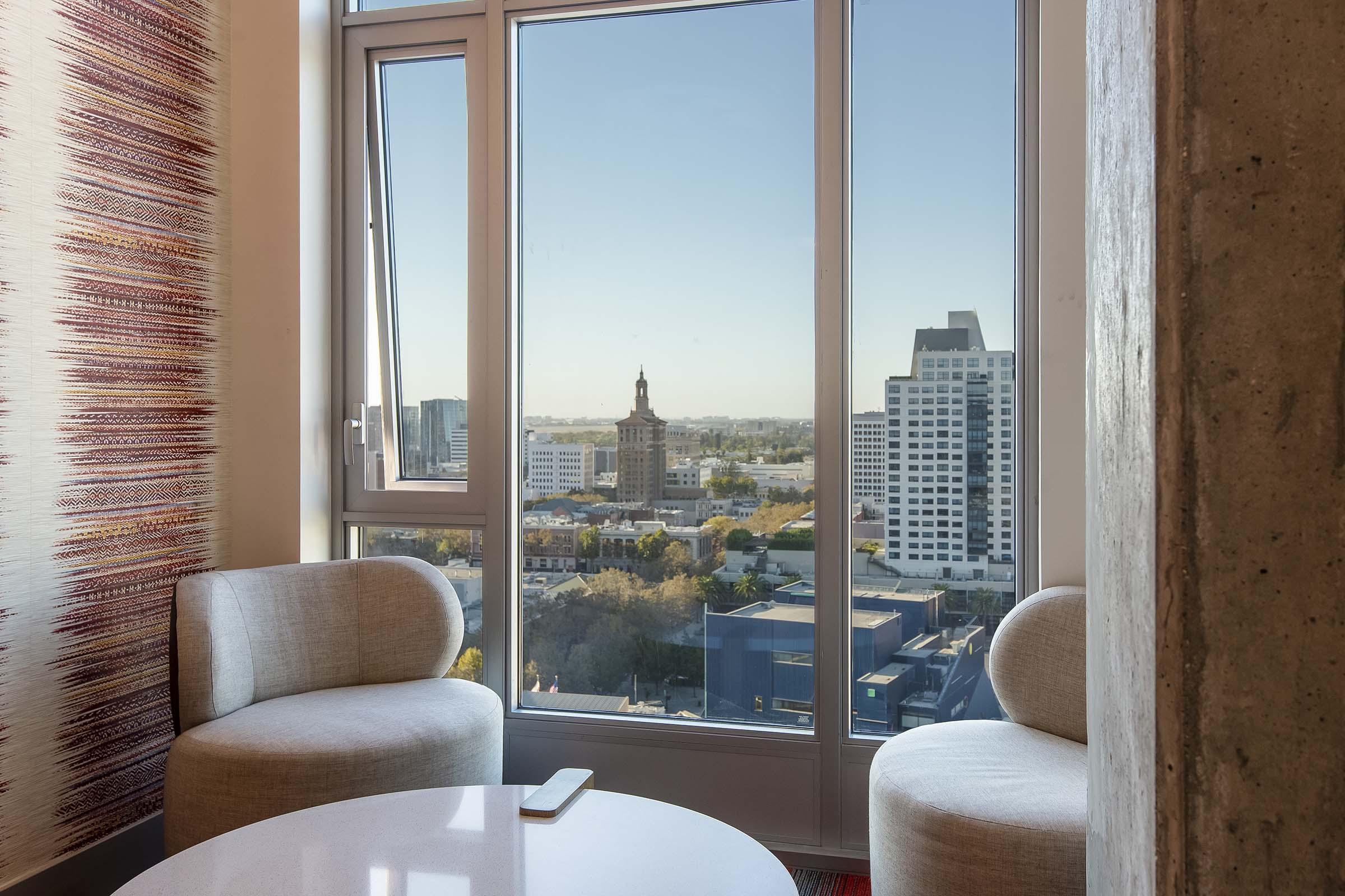 A modern interior view featuring two light-colored chairs beside a round table, overlooking a city skyline through a large window. The scene includes tall buildings and a clear blue sky, suggesting a bright day. Decorative wall elements add a touch of style to the cozy seating area.