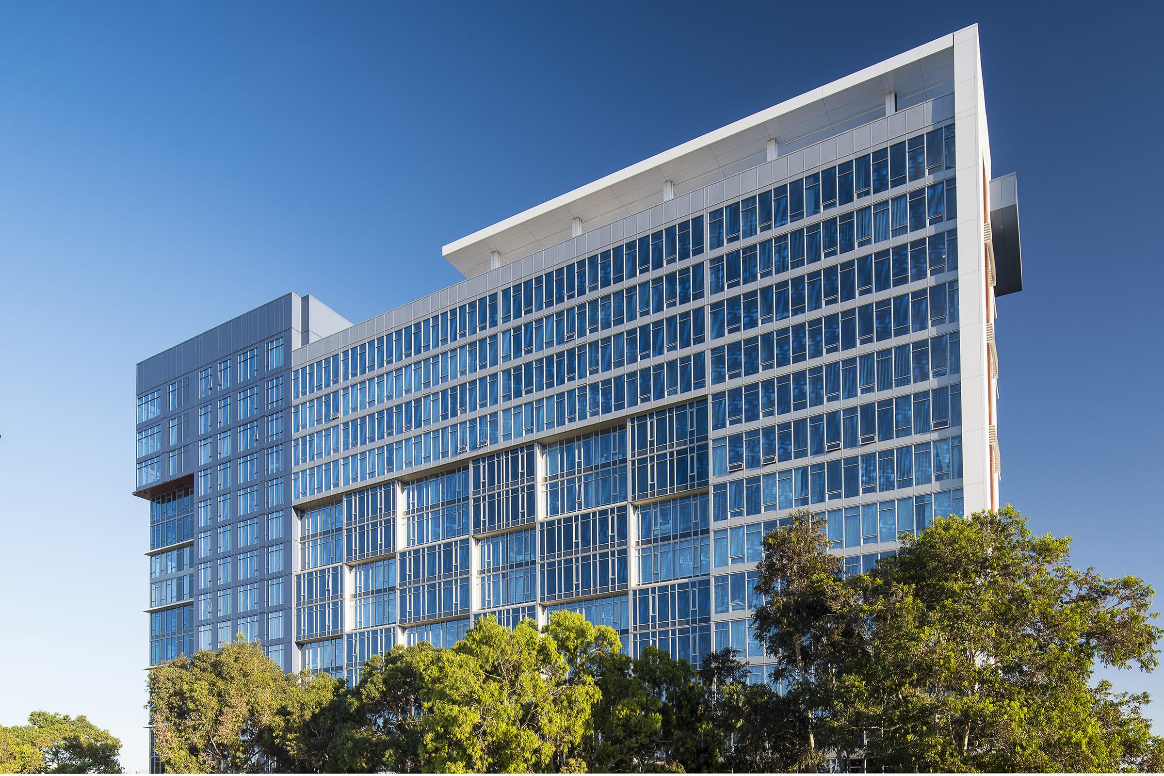 Modern office building with large glass windows and a sleek architectural design, surrounded by greenery under a clear blue sky. The structure features multiple floors and a combination of horizontal and vertical elements, showcasing contemporary urban development.