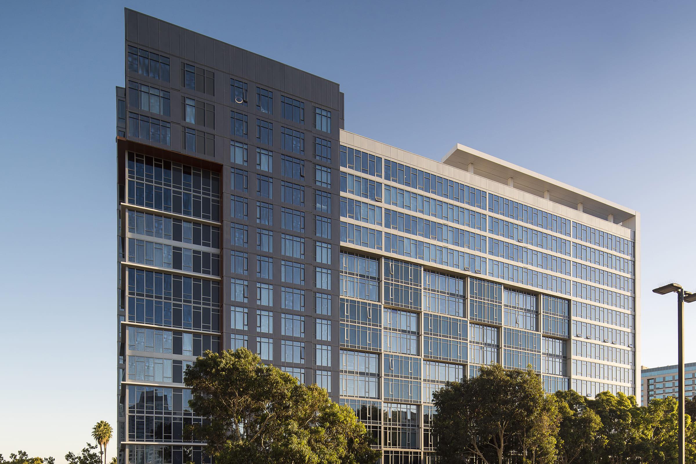 Modern office building with a glass facade, featuring multiple floors and an angular design. Surrounded by trees and clear blue skies, the structure reflects contemporary architectural trends, showcasing large windows that enhance natural light. The setting suggests a professional atmosphere.