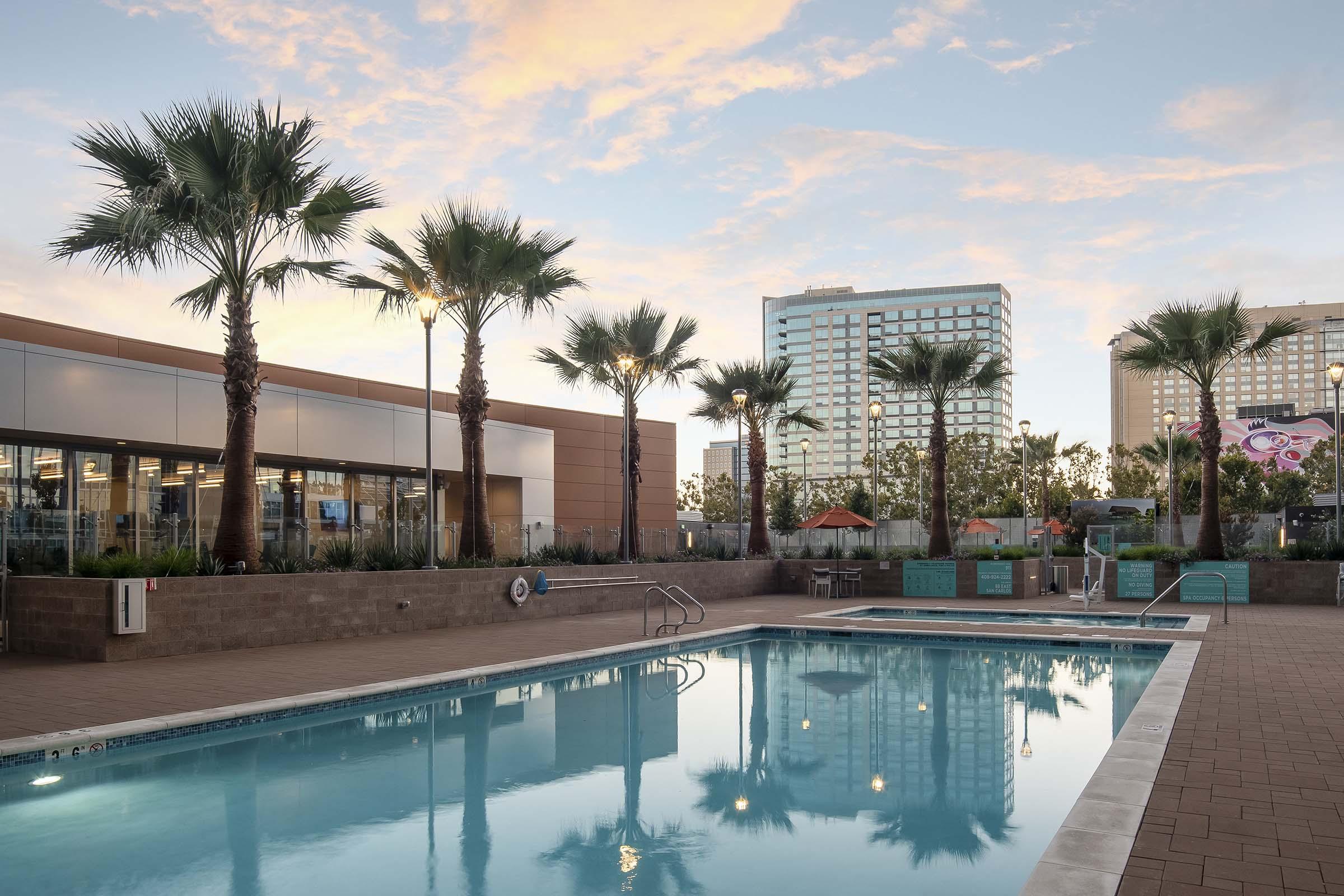 A sparkling outdoor swimming pool surrounded by palm trees, with modern buildings in the background. The sky is painted with soft hues of sunrise or sunset, creating a serene and inviting atmosphere. The pool deck is light-colored brick, enhancing the vibrant scene.