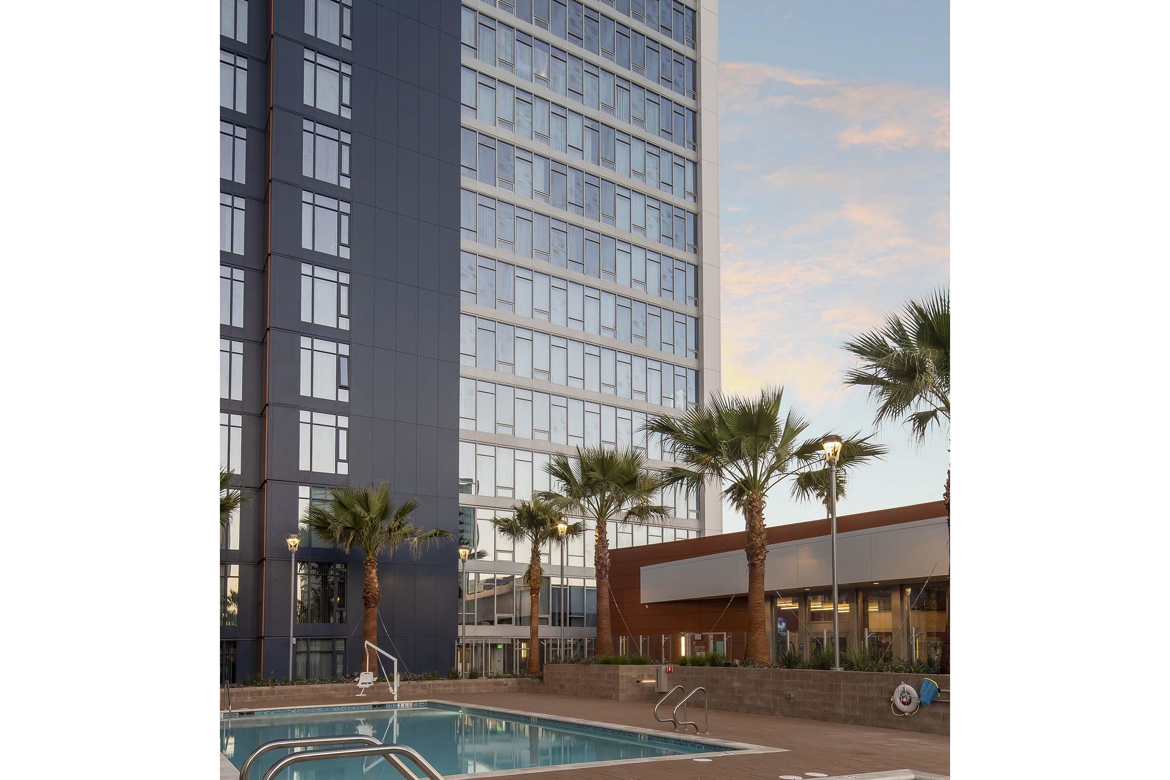 A modern hotel building featuring large glass windows, with a swimming pool in the foreground surrounded by palm trees. The evening sky reflects off the building, creating a serene and inviting atmosphere. The pool area is well-lit, enhancing the overall aesthetic of the scene.