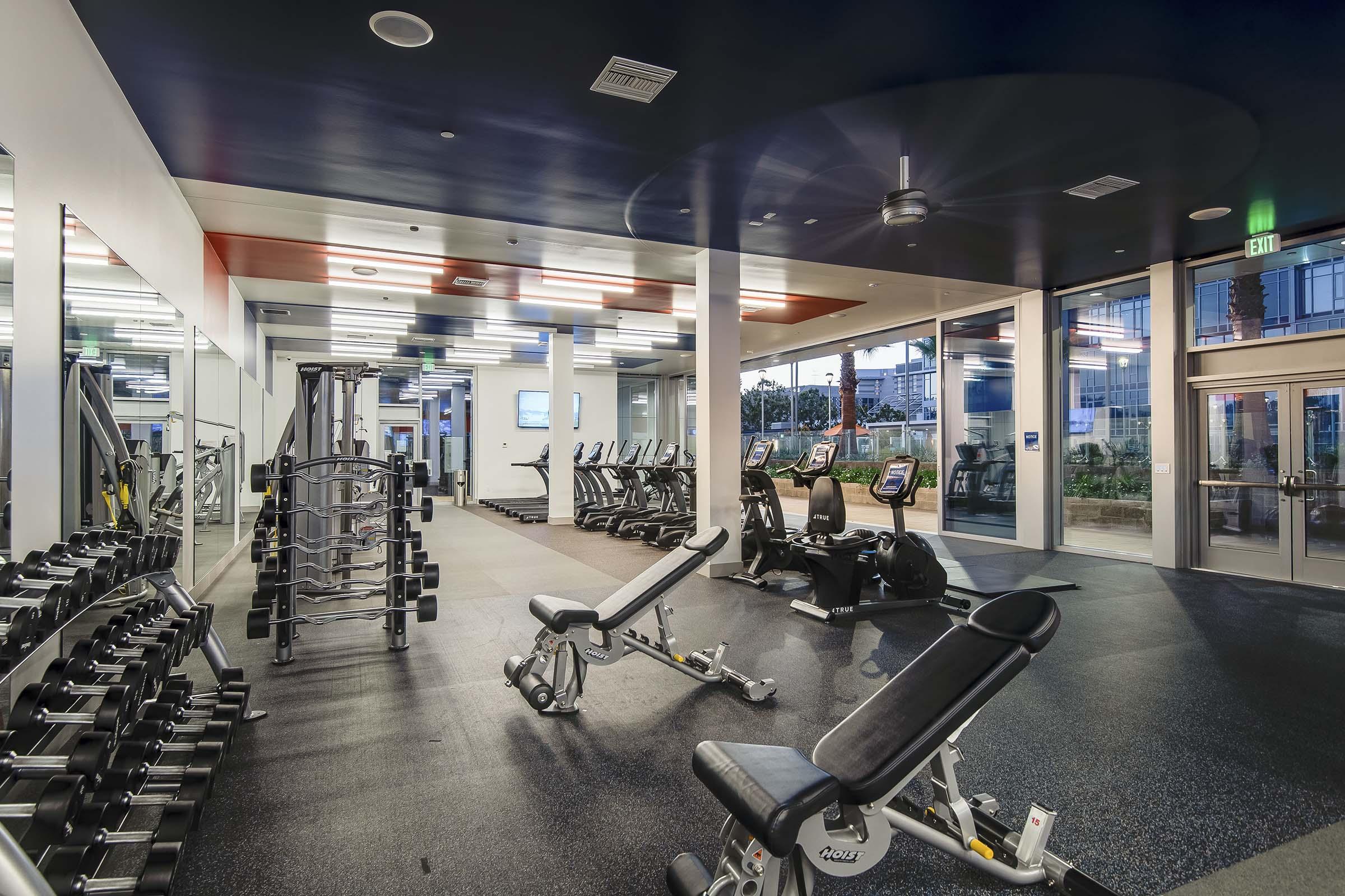 A modern gym interior featuring various workout equipment, including weight benches, free weights, and treadmills. The space has large windows allowing natural light and mirrors lining the walls. The floor is covered in black rubber matting, creating a fitness-friendly environment.