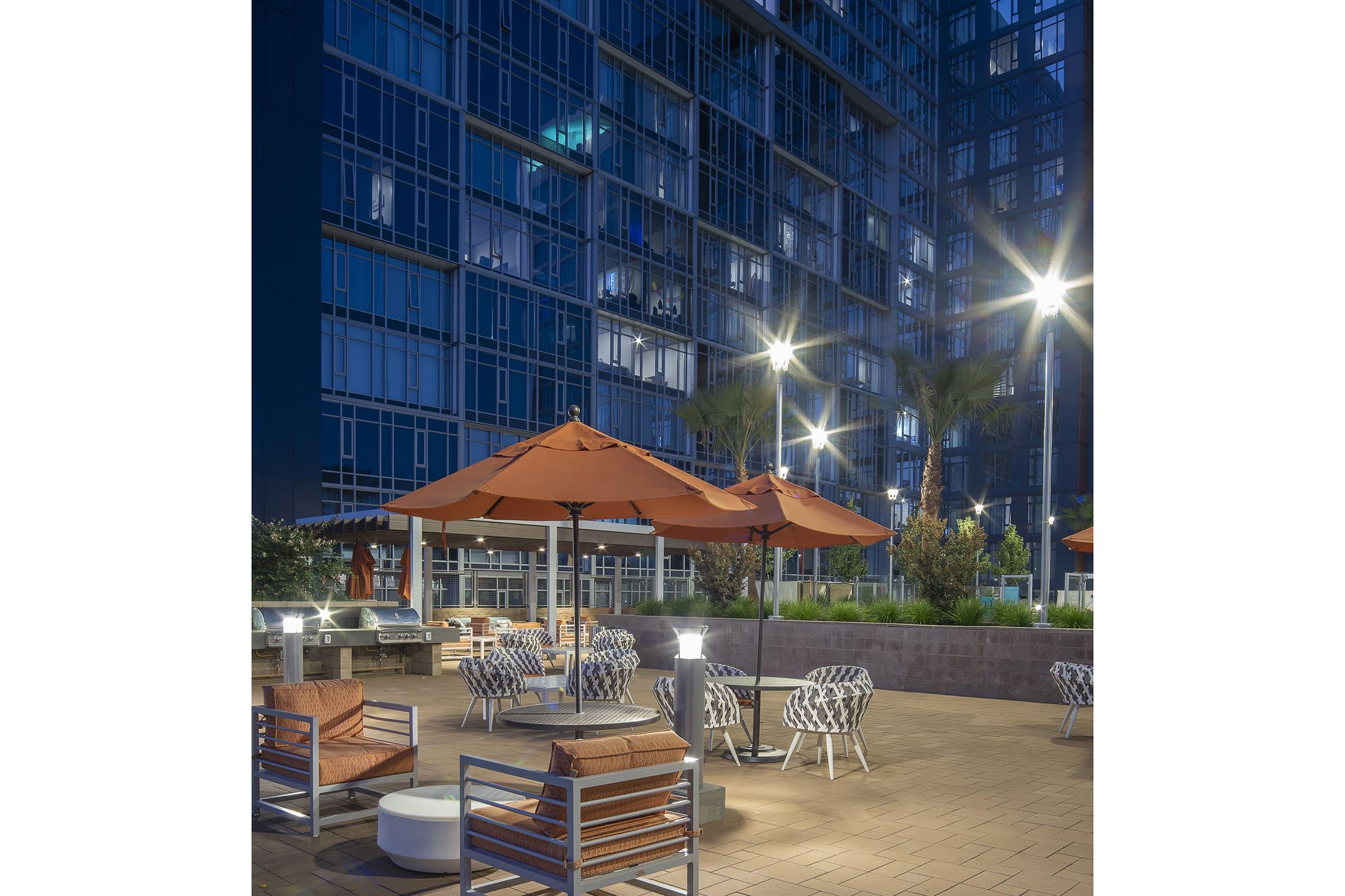 A modern outdoor lounge area featuring stylish seating, orange umbrellas, and well-lit palm trees. The setting is adjacent to a contemporary high-rise building, illuminated at dusk, creating a welcoming ambiance for relaxation and socializing.