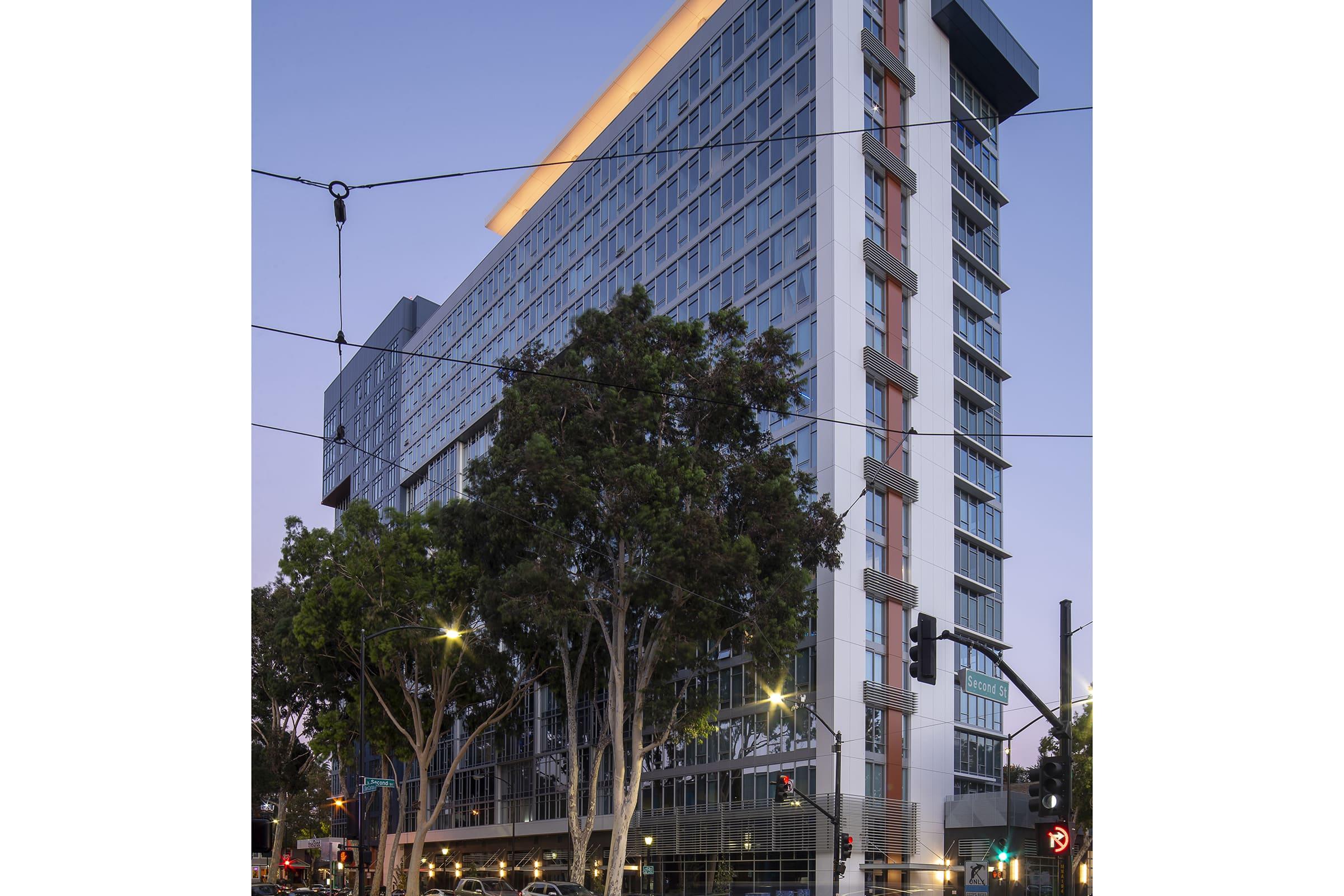 Modern high-rise building at dusk, featuring a mix of glass and concrete architecture. The structure is illuminated with soft lighting along the top edge, surrounded by streetlights and trees, suggesting an urban environment. Power lines and traffic signals are visible in the scene.