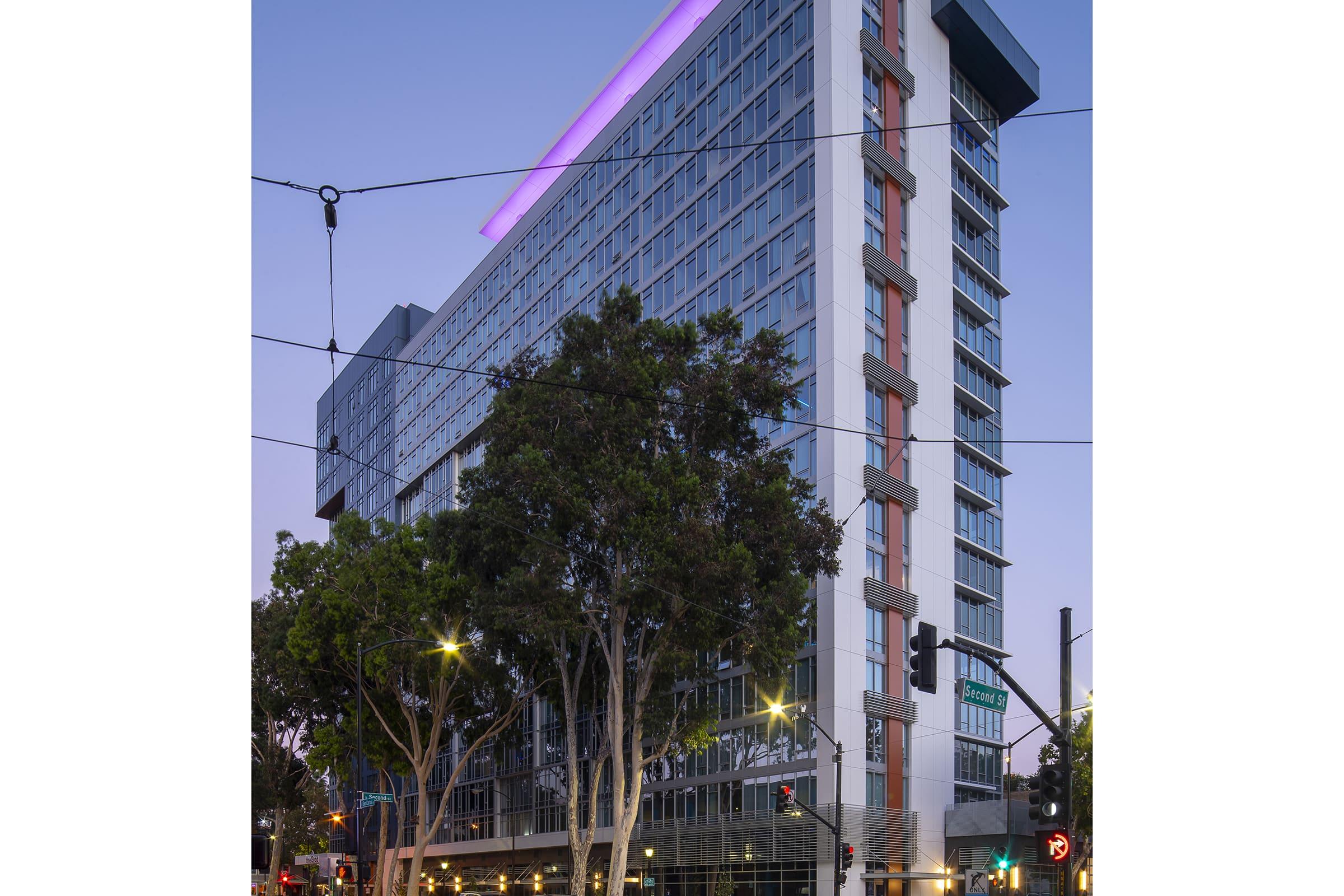 A modern, multi-story hotel building illuminated at dusk, featuring large windows and colorful lighting on the roof. Surrounding the hotel are trees and traffic signals, with streetlights casting a warm glow. The setting suggests a vibrant urban area.