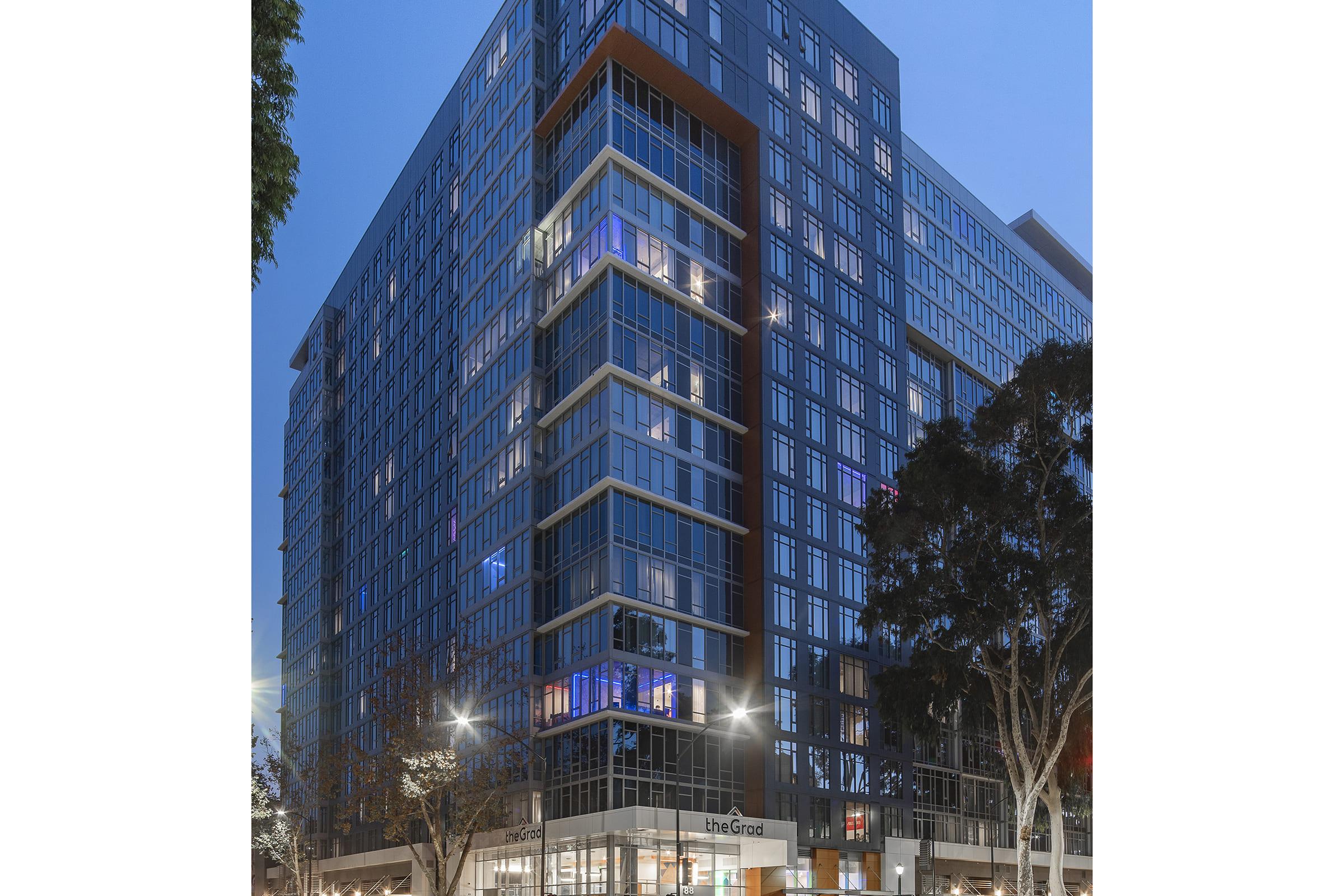 A modern multi-story residential building with large glass windows, illuminated at dusk. The structure features a sleek, contemporary design and a welcoming entrance. Surrounding trees and streetlights add to the urban atmosphere, showcasing a vibrant city scene.