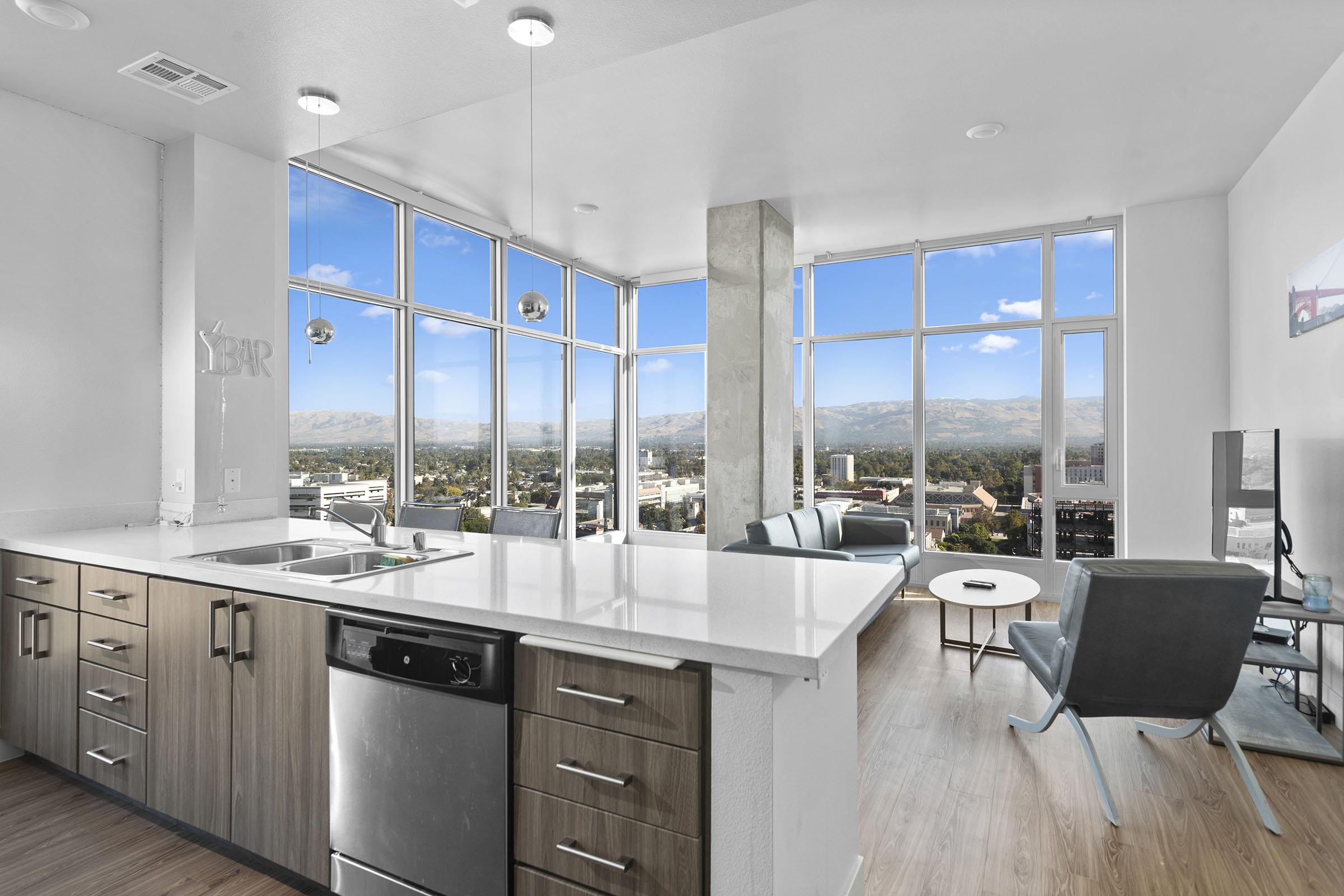 A modern kitchen and living area with large windows offering a panoramic view of mountains and a cityscape. The kitchen features a stainless steel sink and appliances, sleek cabinetry, and a stylish countertop. The living area has comfortable seating and a coffee table, all illuminated by natural light.