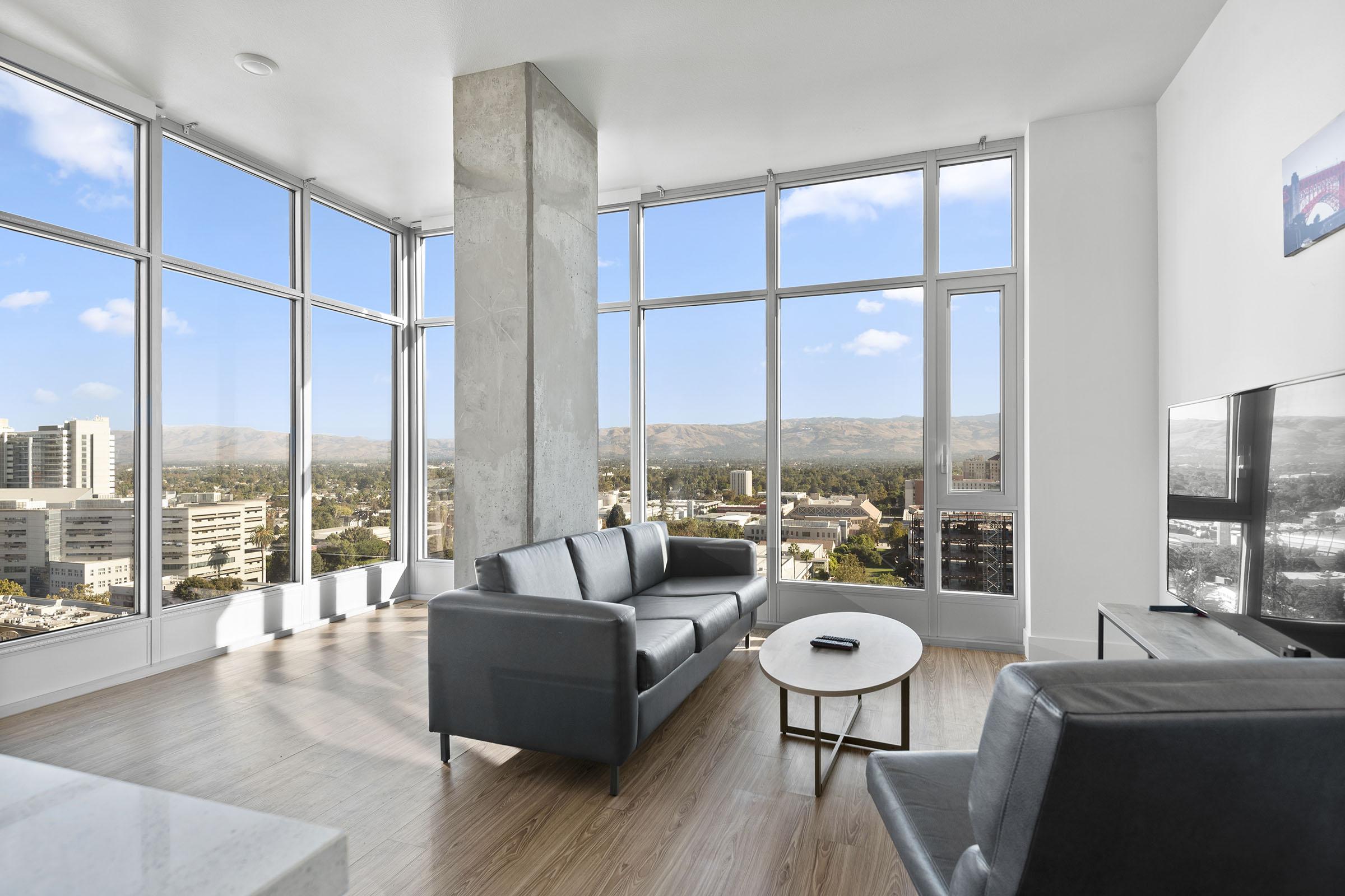 A modern living room with large windows showcasing a panoramic view of mountains and urban landscape. The space features stylish furniture, including a gray sofa, an armchair, and a circular coffee table, bathed in natural light. The walls are white, and there is a flat-screen TV in the corner.