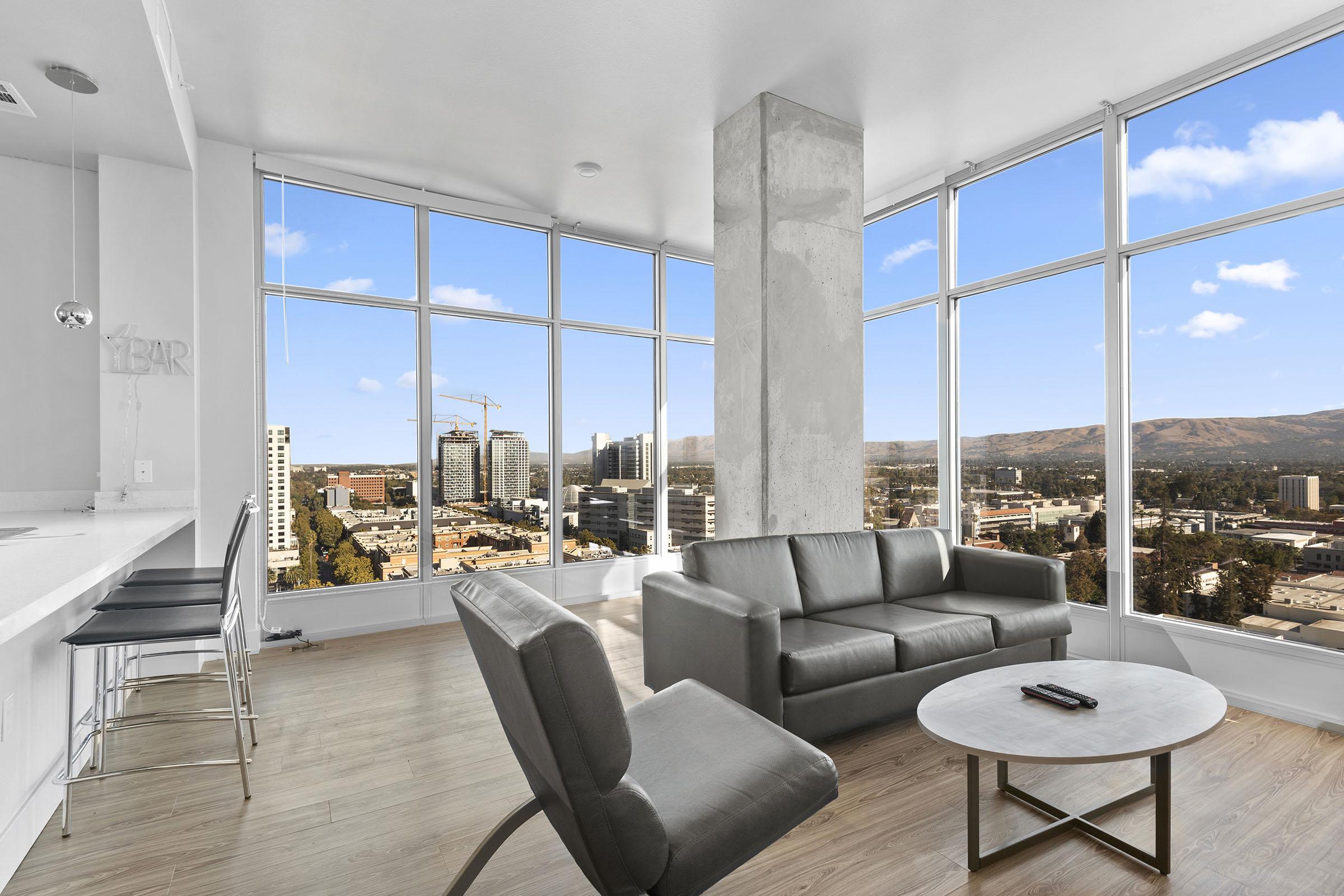 Modern apartment interior featuring large windows with city and mountain views. The space is furnished with a gray couch, an armchair, and a round coffee table. Light wood flooring complements the contemporary design. The bright room has an open feel, showcasing urban scenery outside.