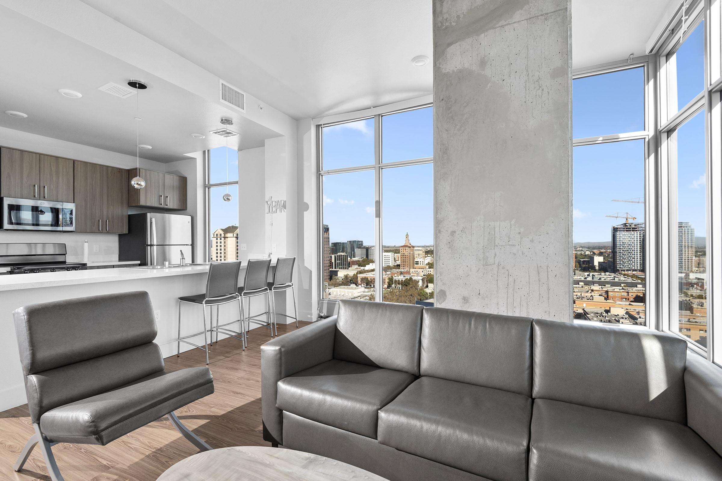 A modern living room with large windows showcasing a city skyline. The space features a gray leather couch, a round coffee table, and sleek bar stools at a kitchen counter. Sunlight streams in, highlighting the contemporary decor and concrete accents.