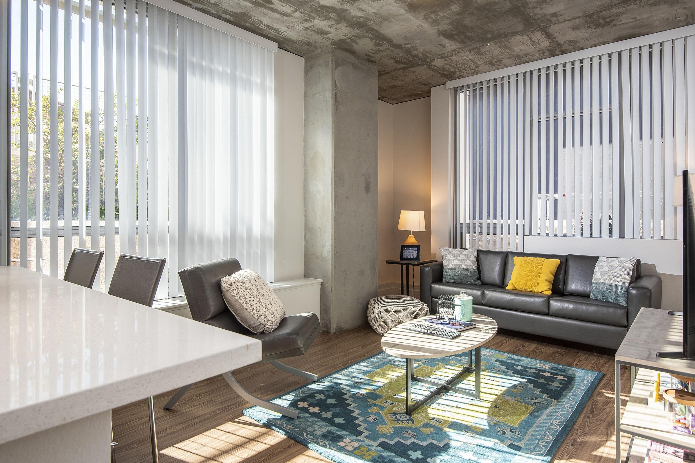 A modern living room featuring large windows with vertical blinds, a gray sofa with colorful cushions, a round coffee table on a patterned rug, a floor lamp, and minimalist decor. Sunlight filters in, creating a bright and inviting atmosphere.