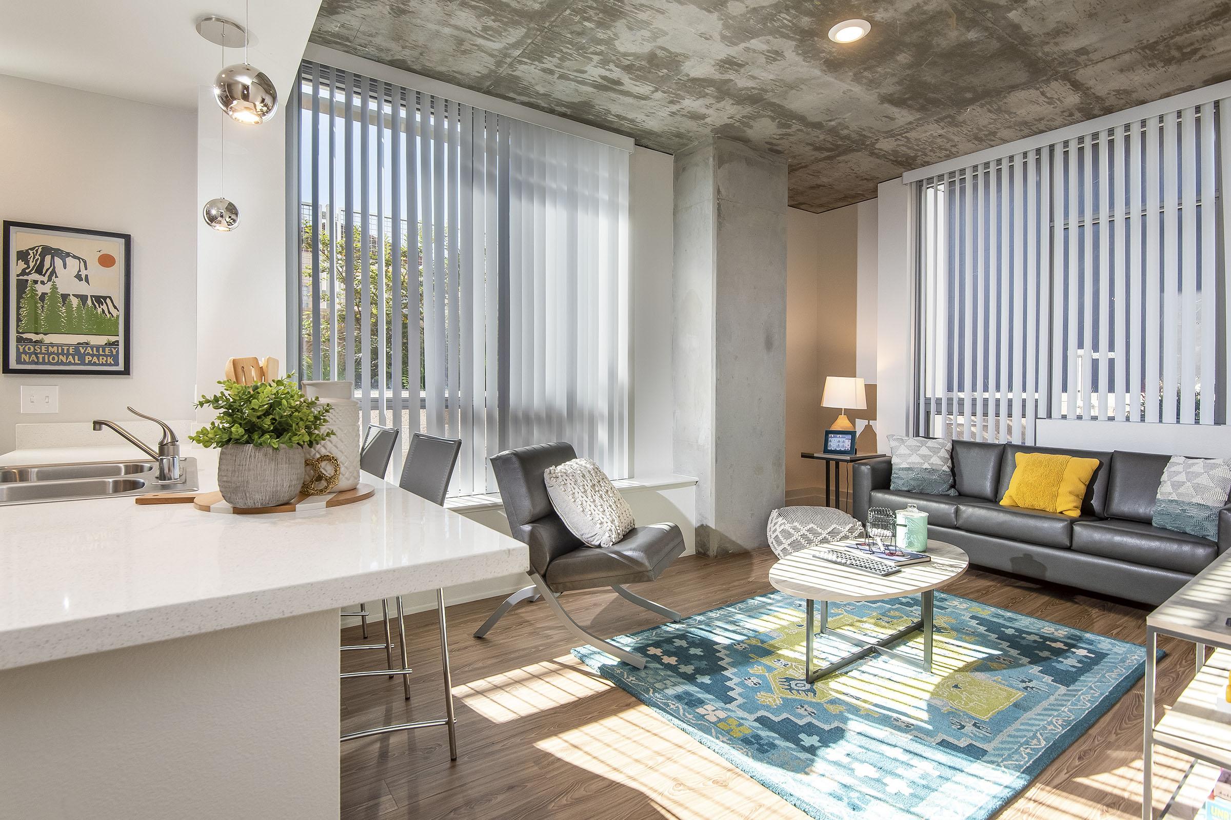 Modern living room with large windows and vertical blinds, featuring a sleek sofa adorned with colorful cushions, a coffee table, and a bright area rug. The kitchen area includes a white countertop and bar stools, while light fixtures hang from the ceiling, complemented by a houseplant. The walls have a contemporary, industrial finish.