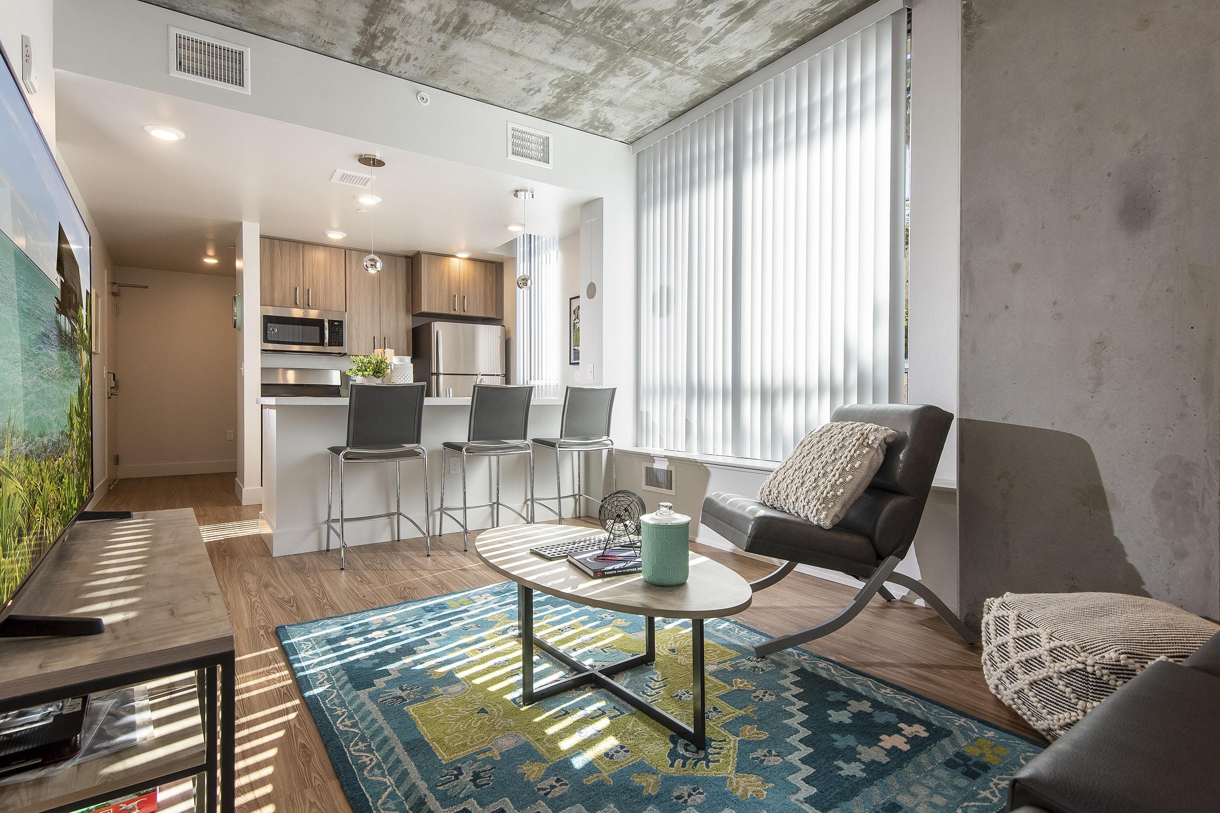 Modern living room in a stylish apartment featuring a concrete wall and ceiling. A cozy seating area includes a sleek chair and a coffee table on a decorative rug. In the background, there is an open kitchen with bar stools and stainless steel appliances, along with large windows covered by vertical blinds allowing natural light.