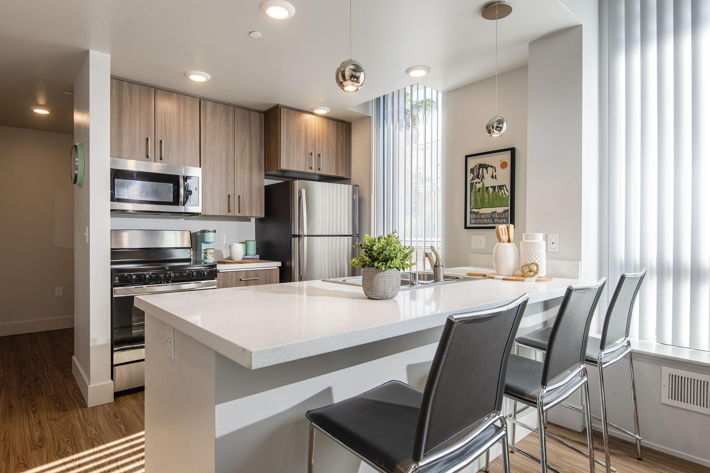 Modern kitchen featuring a spacious island with bar stools, stainless steel appliances including a microwave and fridge, and light wood cabinetry. Large windows with vertical blinds allow natural light to fill the space, creating a bright and inviting atmosphere. A small plant adds a touch of greenery to the countertop.