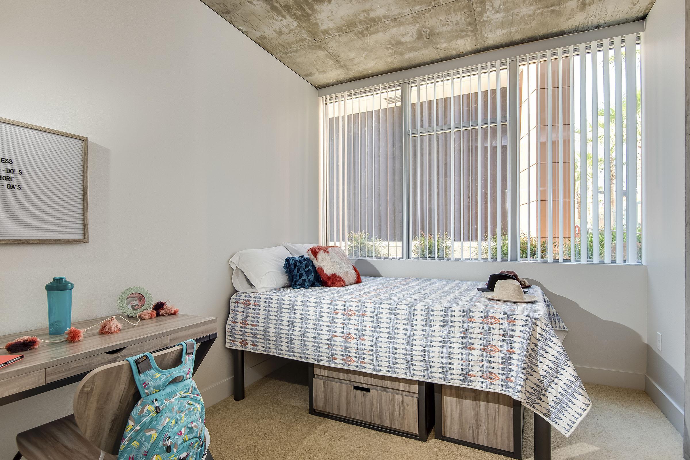 A neatly arranged bedroom featuring a single bed with decorative pillows, a small desk with a water bottle and various items, and a backpack. Large windows provide natural light and reveal a view of outdoor greenery. The room's walls are light-colored, and the flooring is carpeted.