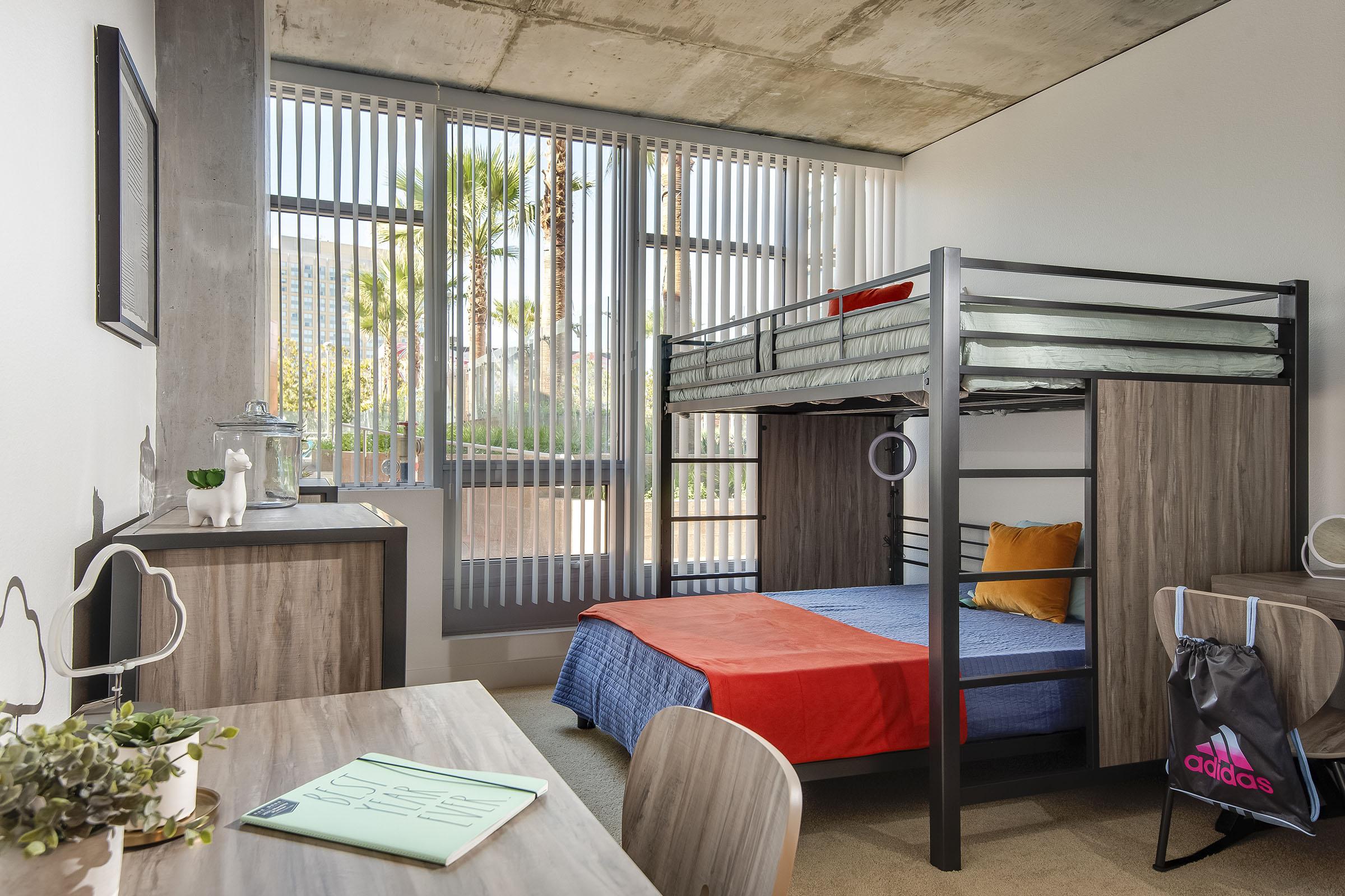 A modern bedroom featuring a lofted bed with a green mattress and an orange blanket on the lower level. The room has large windows with vertical blinds, allowing natural light. There's a wooden desk with a notepad, a small plant, and a closet. A chair and a bag with the Adidas logo are visible.