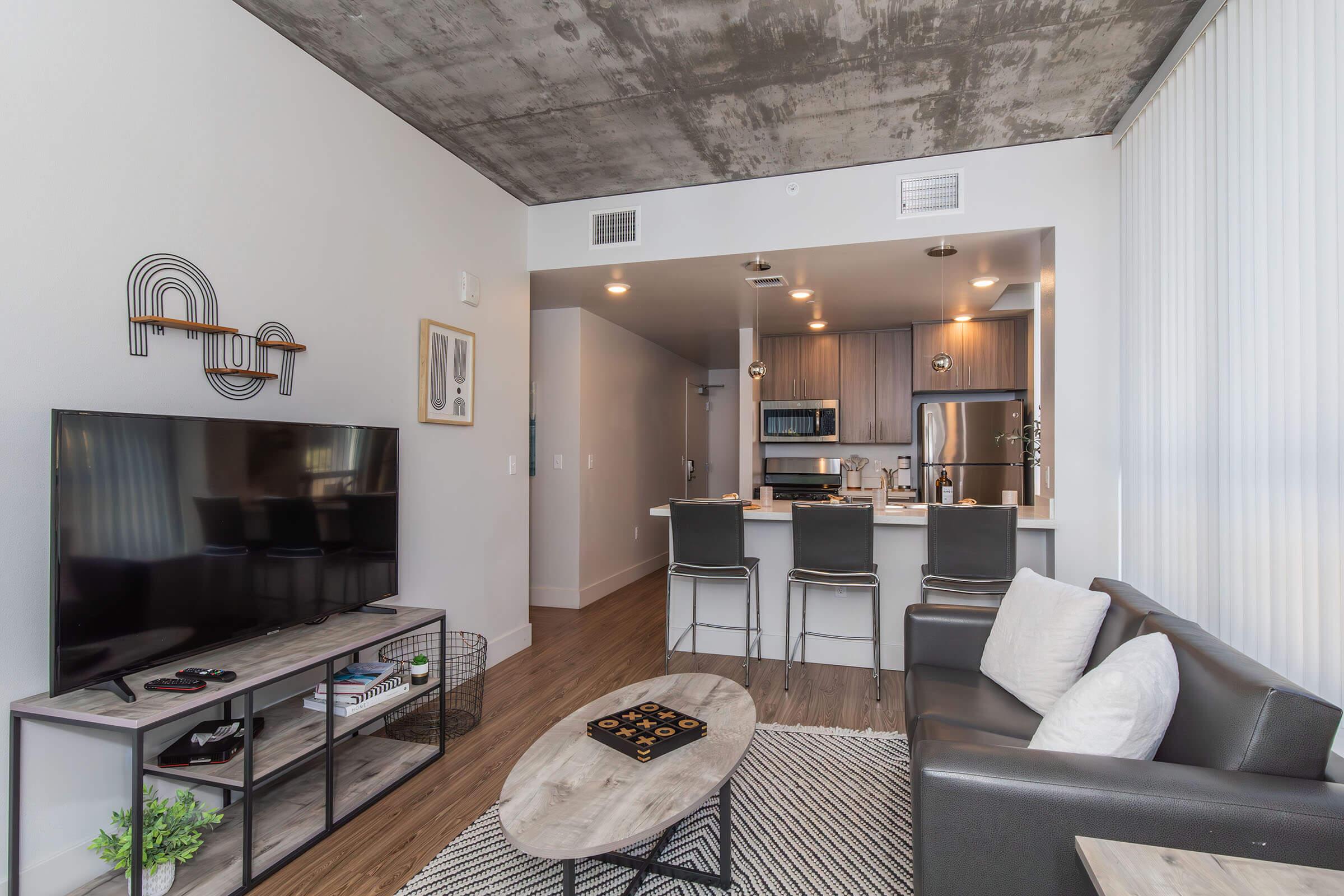 Modern living room featuring a flat-screen TV on a stand, a coffee table, and a sectional sofa. The space has a concrete ceiling and is open to a kitchen with bar seating. Light wood flooring complements the neutral color scheme. Decorative wall art adds a touch of style to the contemporary atmosphere.